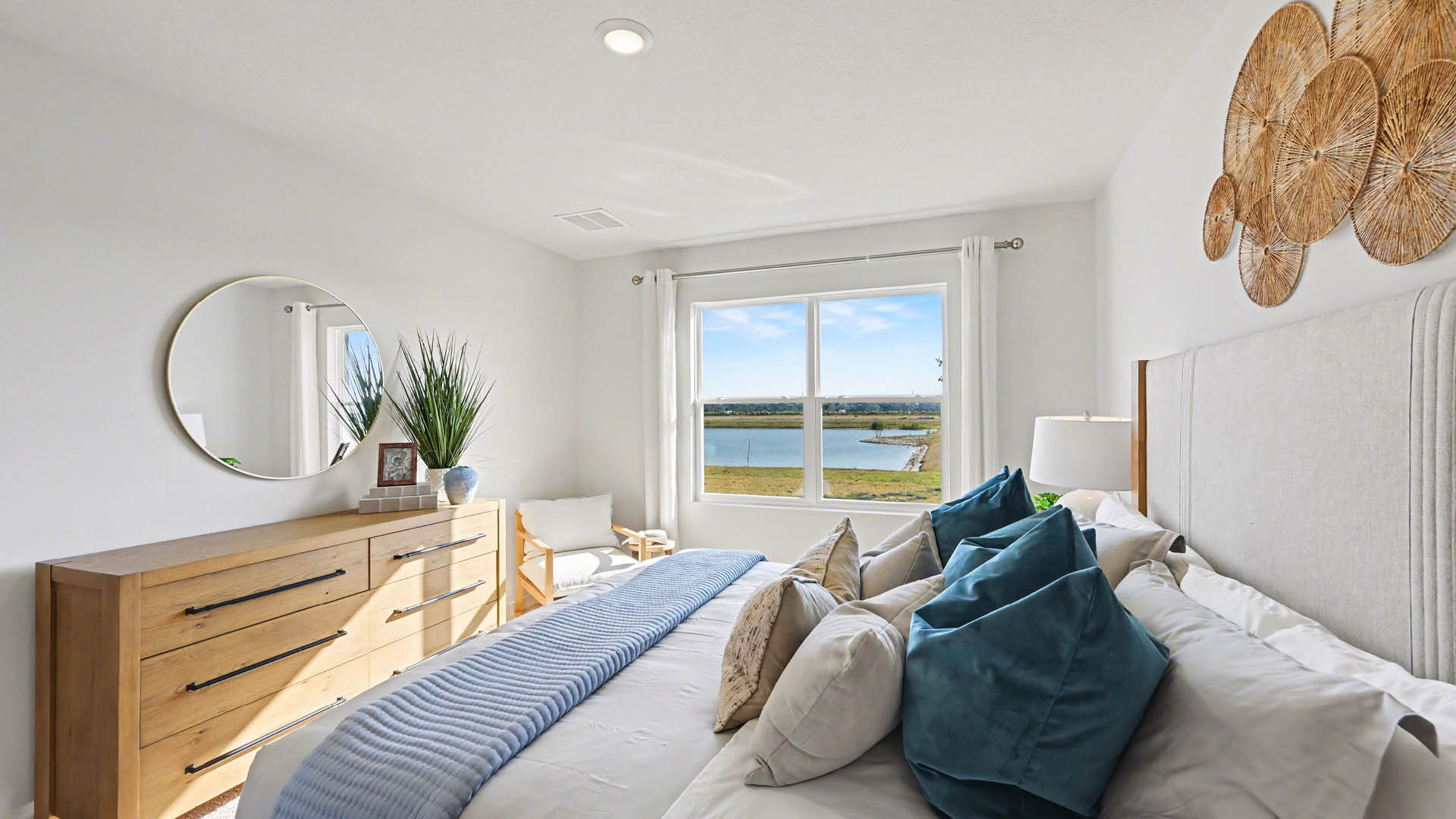 A bedroom with a large bed, a window, and a mirror. The bed is covered with pillows and blankets. The window overlooks a body of water.