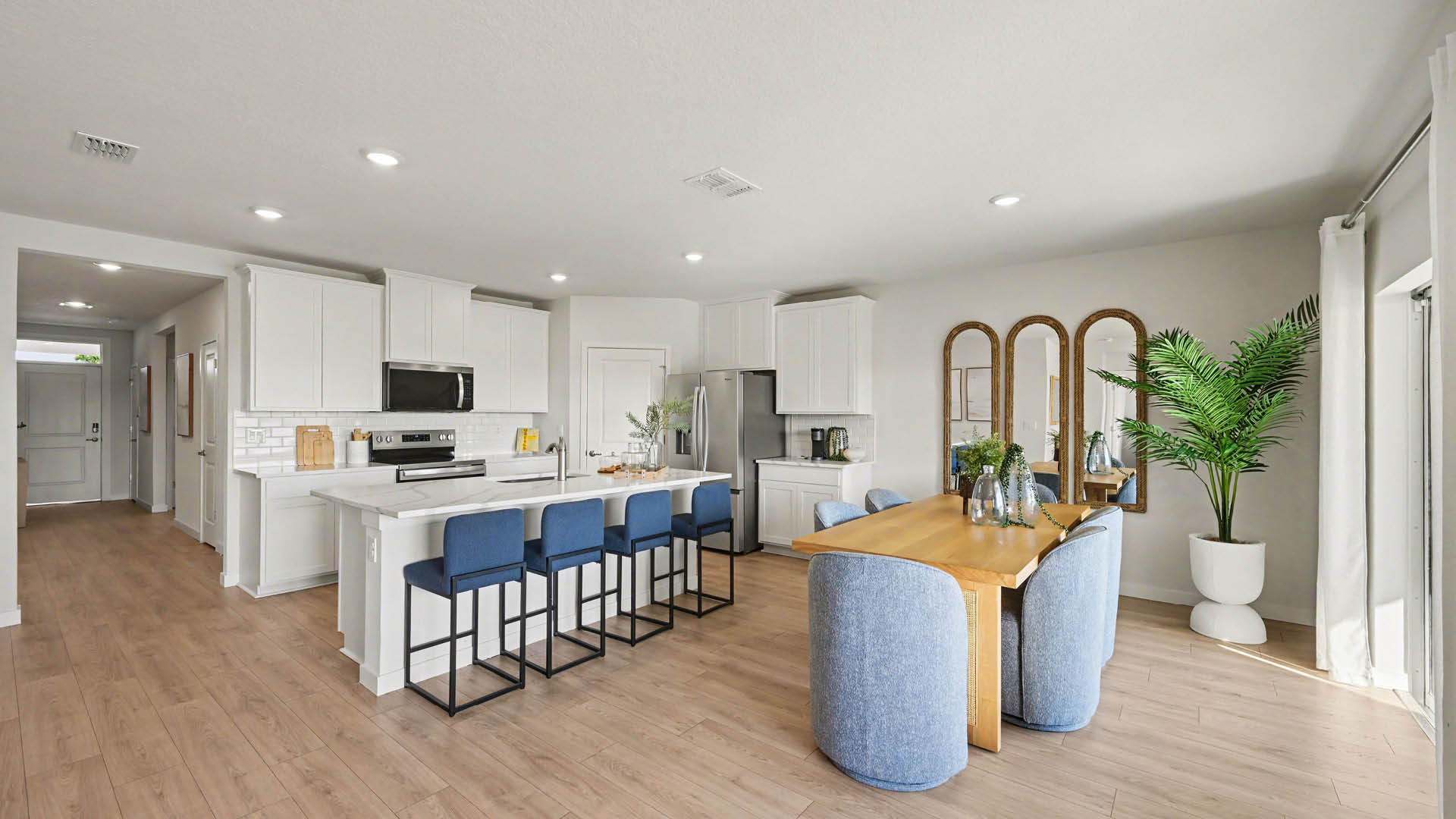Modern kitchen with white cabinetry, stainless steel appliances, and a large island with seating. A cozy dining area with large windows and a welcoming entryway are visible.