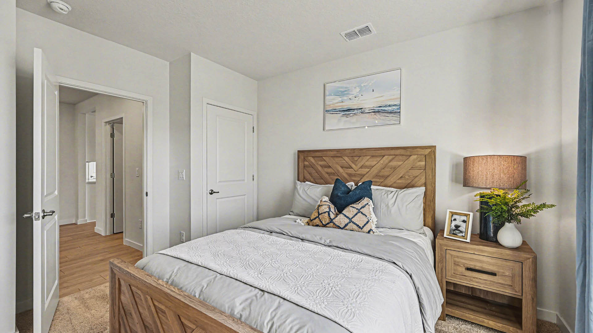 A bedroom with a bed, nightstand, and lamp. The room has white walls,  beige carpet, a closed closet door and a hallway leading to another area.