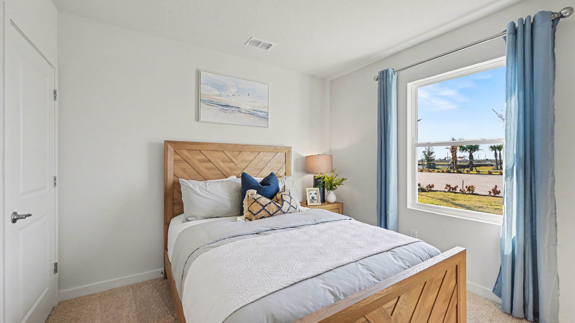 A bedroom with a bed, a window, and a painting on the wall. The room has white walls and beige carpet. A closed door is visible.