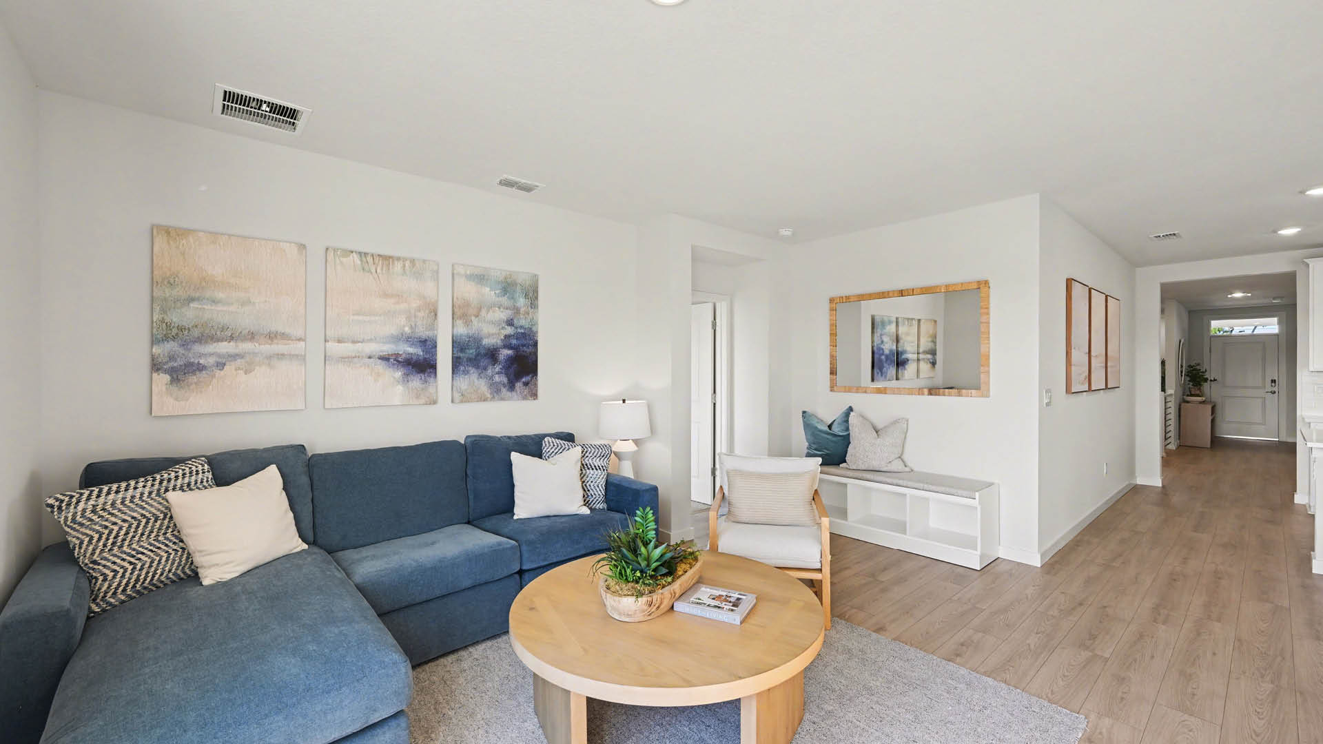 Spacious living room with plush seating area and rug. The room has large windows, and minimalistic walls, allowing natural light to enter. An entryway hallway leading to the front door is visible.