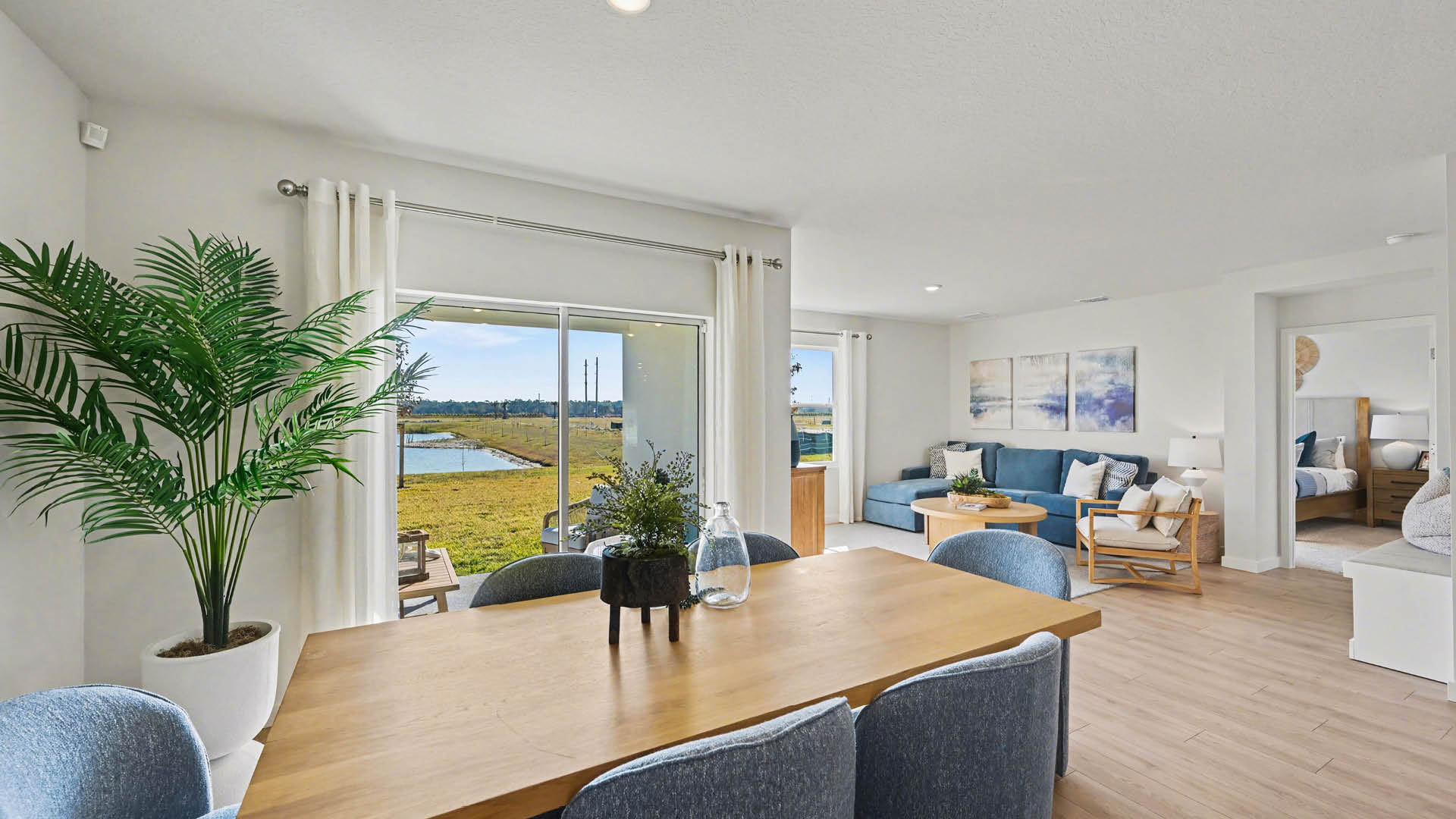 Bright dining area featuring large windows and sliding glass doors opening to a green outdoor area. The living room and a doorway to a bedroom are visible.