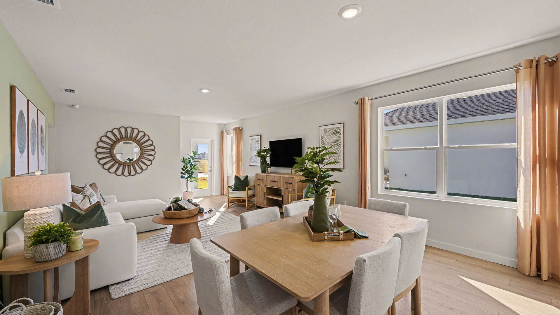 A bright dining area featuring large windows and a door leading to a green outdoor area. The living room with plank flooring is visible.