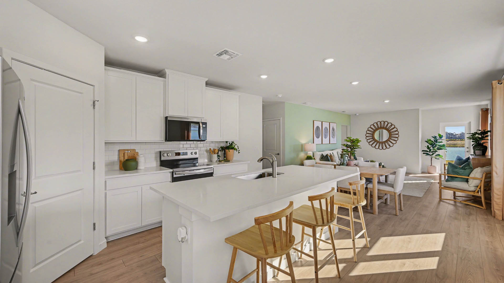 A large, clean kitchen with a center island with seating and stainless steel appliances. A dining area and living room are also visible.