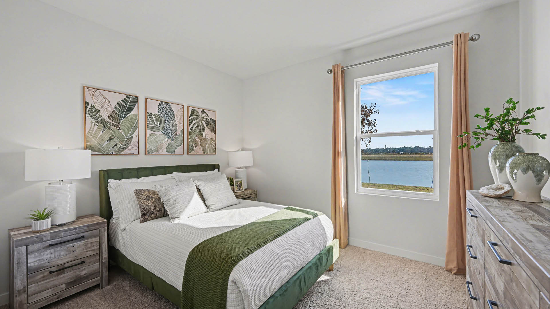 A bedroom with a bed, a window, and a painting on the wall. The room has white walls and beige carpet. The window looks out onto beautiful water.