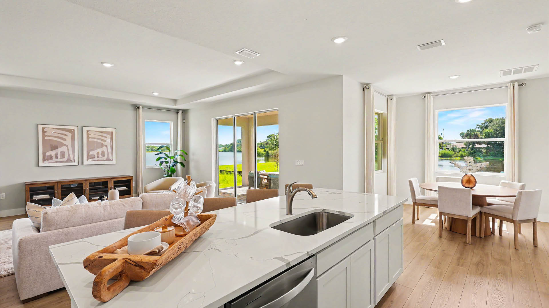 A modern open-concept kitchen and living area features a countertop island with a sink, a dining table by large windows, and a view of greenery and water outside.