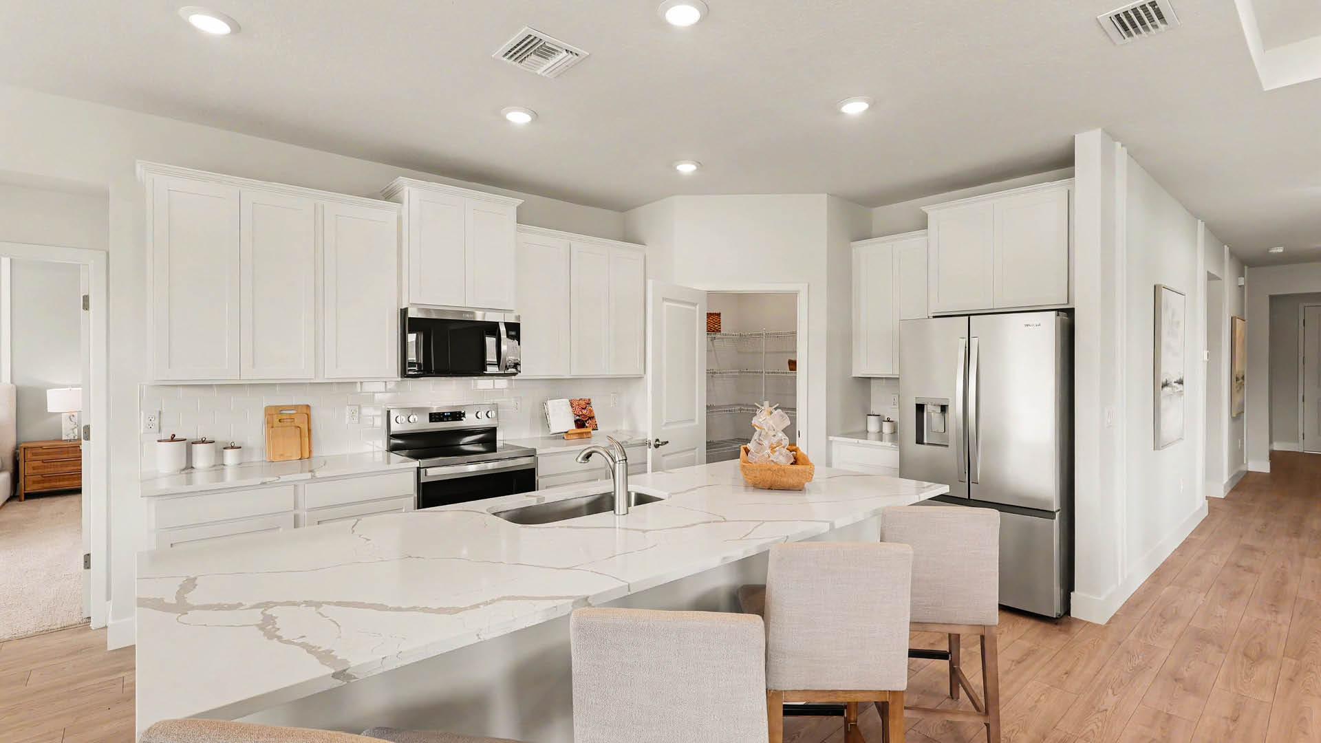 A modern kitchen features white cabinetry, stainless steel appliances, an island with four beige upholstered chairs, and recessed ceiling lights.