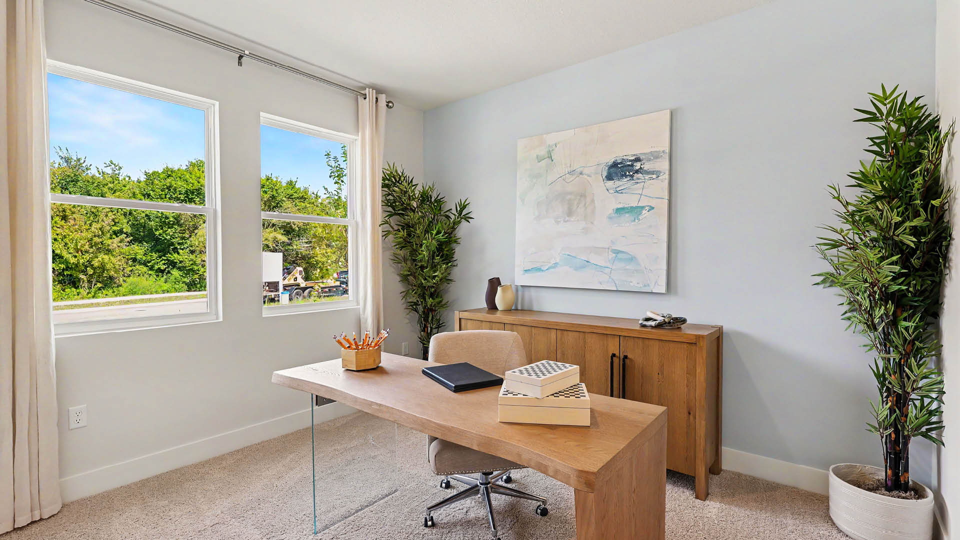 A modern home office with a wooden desk, office chair, two potted plants, a sideboard with decorative items, abstract wall art, and two large windows showing greenery outside.
