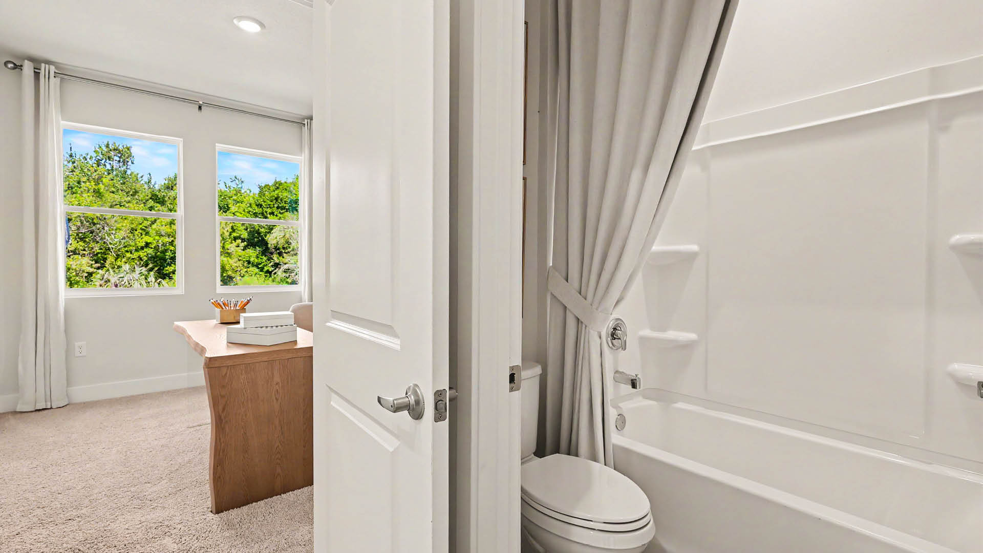 A bright room with a wooden desk and large windows opens into a small bathroom featuring a bathtub with a shower curtain and a toilet.