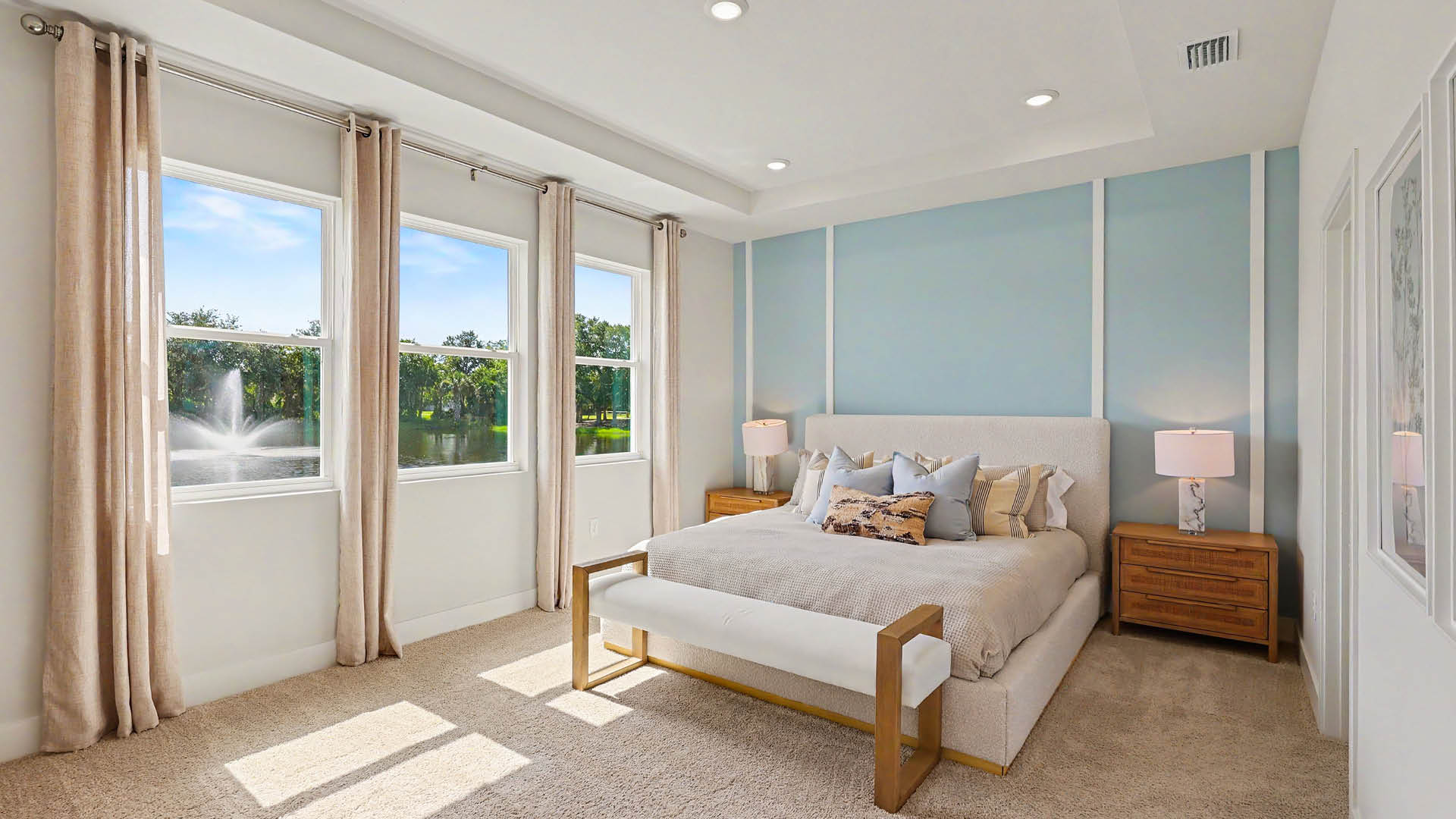 A modern bedroom features a large bed with multiple pillows, two wooden nightstands with lamps, beige curtains framing three windows overlooking a garden with a fountain, and soft carpeted flooring.