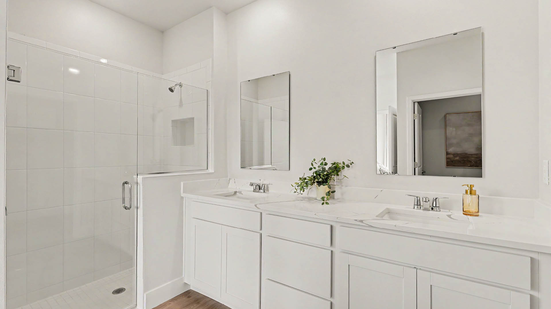 A modern bathroom features a white double vanity with two mirrors, a small plant, soap dispenser, and a glass-enclosed shower with white tiled walls.