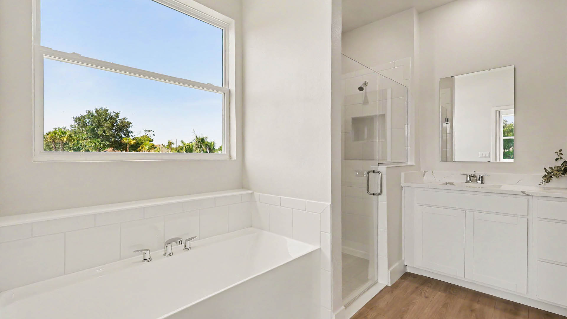 A bright bathroom features a white bathtub beneath a window, a glass-enclosed shower, and a white vanity with a mirror and faucet.
