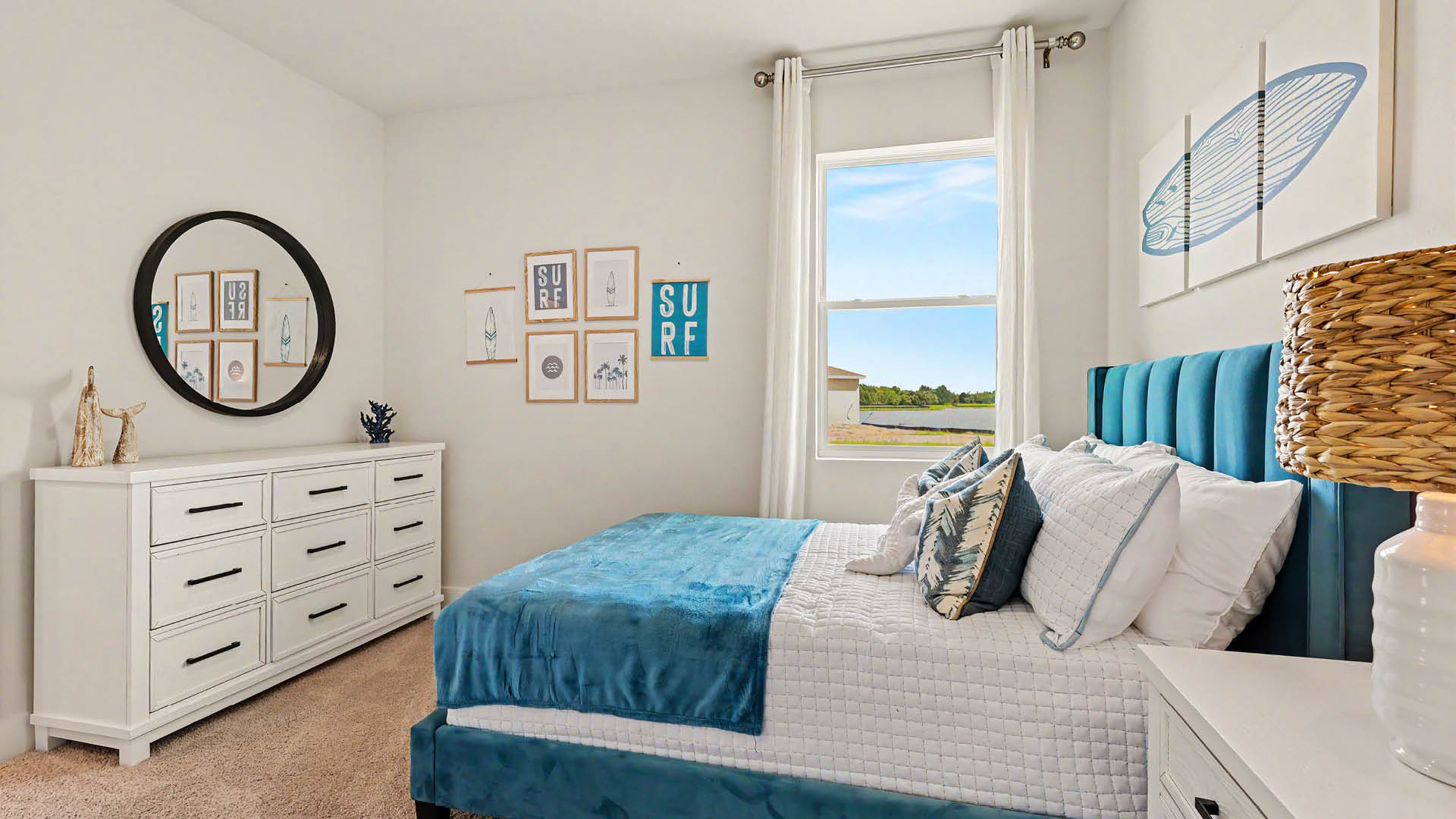 A bright bedroom features a blue upholstered bed with white and blue bedding, white furniture including a dresser and nightstand, wall art with a surf theme, a round mirror, and a window showing an outdoor view.