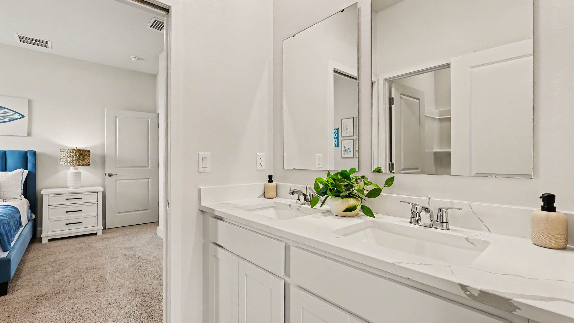 A modern bathroom with a double sink vanity featuring white cabinets, two mirrors, and a small plant, with a view into a carpeted bedroom with a blue bed and white nightstand.