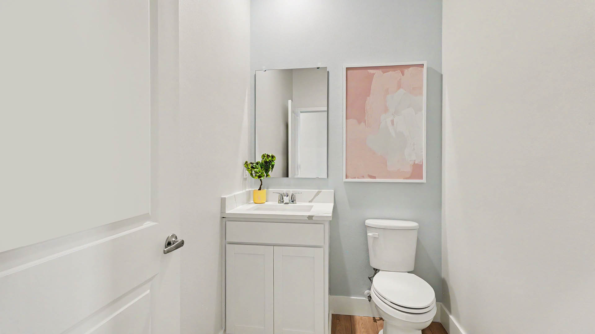 A small bathroom features a white vanity with a mirror, a toilet, a pastel abstract painting on the wall, and a small plant in a yellow pot on the countertop.