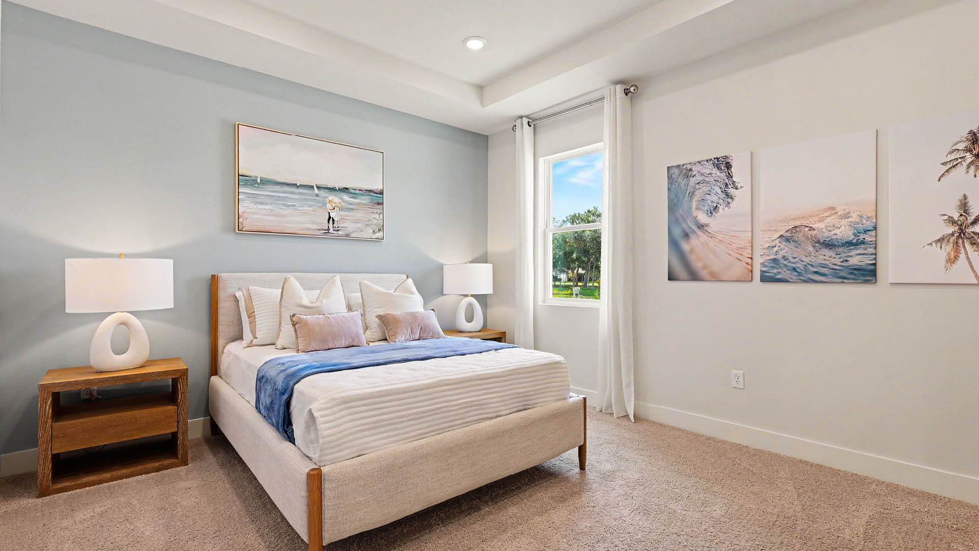 A modern bedroom features a neatly made bed with white and blue bedding, two matching bedside tables with white lamps, light blue and white walls, three ocean-themed paintings, and a window with white curtains showing greenery outside.