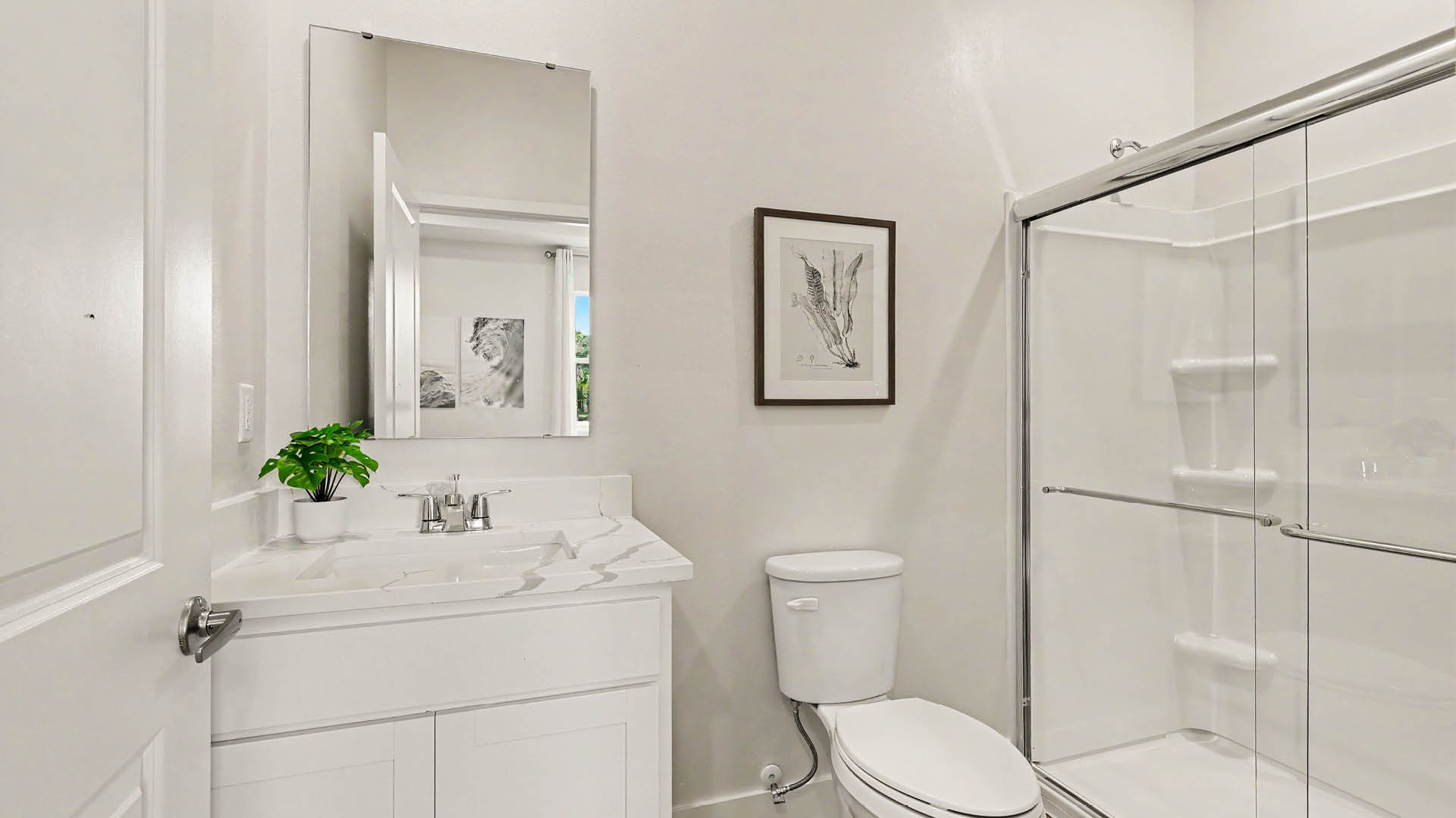 A clean bathroom features a white vanity with a small green plant, a rectangular mirror, a toilet, and a glass-enclosed shower.