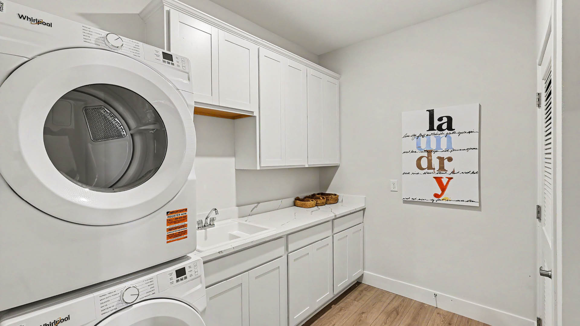 A laundry room with stacked white Whirlpool washer and dryer units, white cabinets, a sink, and a wall decoration spelling out the word laundry.