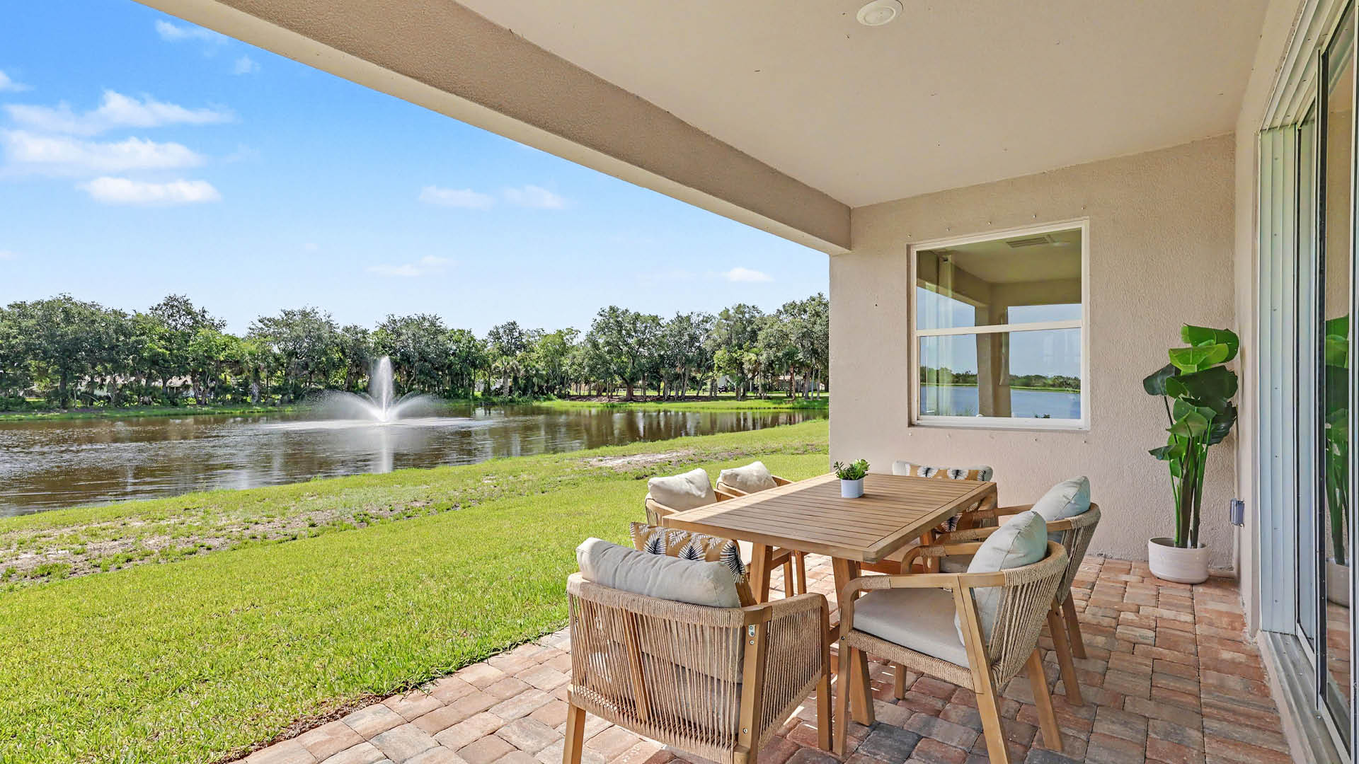 A covered patio with a wooden table and cushioned chairs overlooks a grassy area and a lake with a water fountain under a clear blue sky.