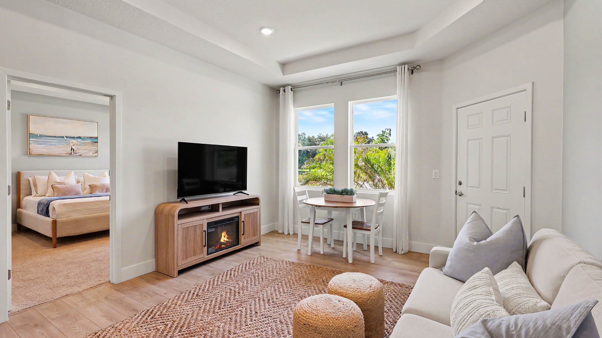 A modern living room with a beige sofa, two round ottomans, a wooden TV stand with a flat-screen TV and electric fireplace, a small dining table with chairs near a window, and a view into a bedroom with a bed and wall art.