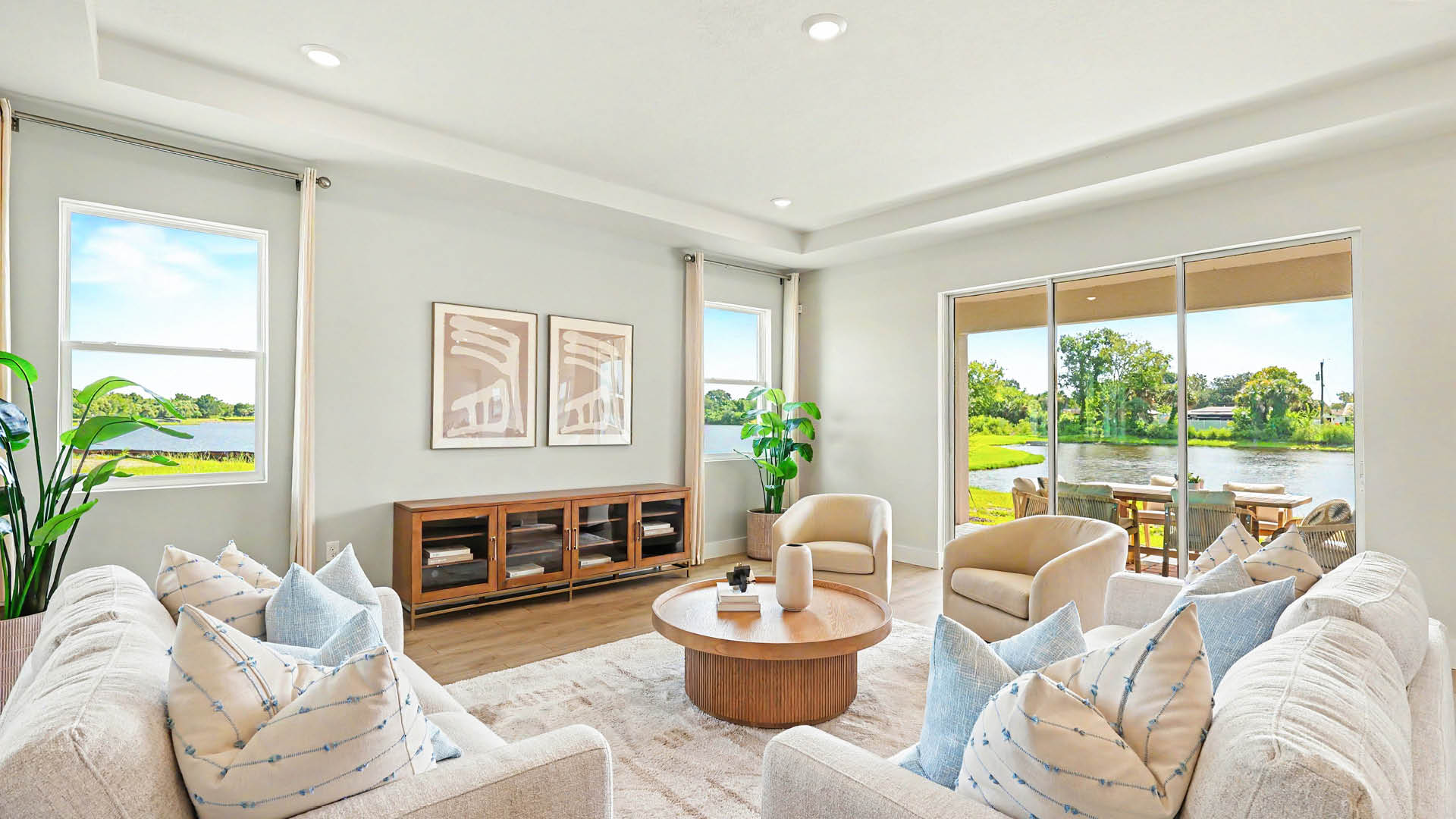 A bright and modern living room features two beige sofas with patterned blue pillows, two armchairs, a round wooden coffee table, a wooden sideboard with framed artwork above, large windows, and sliding glass doors overlooking a green outdoor area with water.