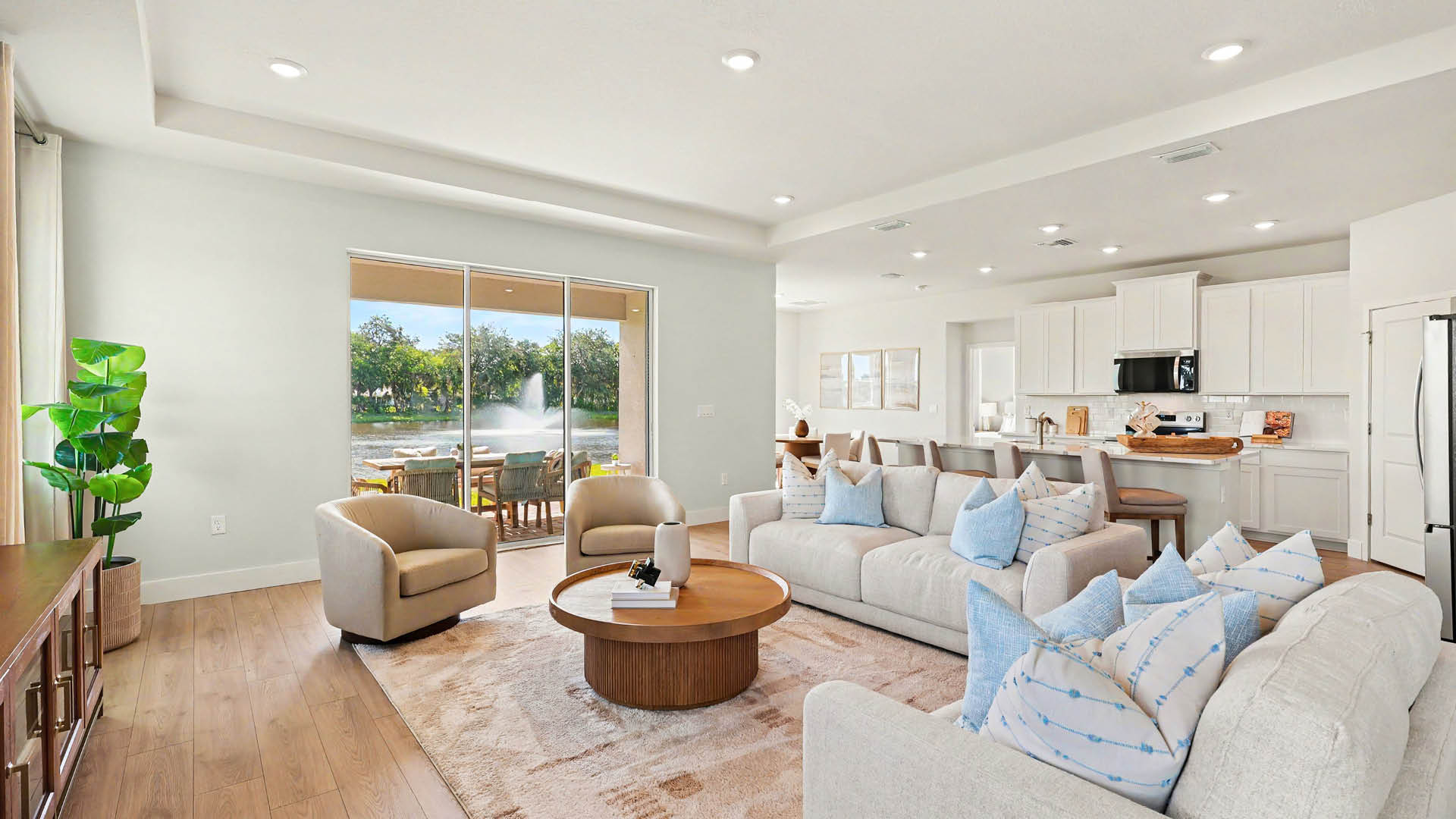 A modern living room with light-colored sofas adorned with blue and white pillows, a round wooden coffee table, two armchairs, a large rug, and a view of a patio and water fountain through sliding glass doors. A kitchen is also visible.