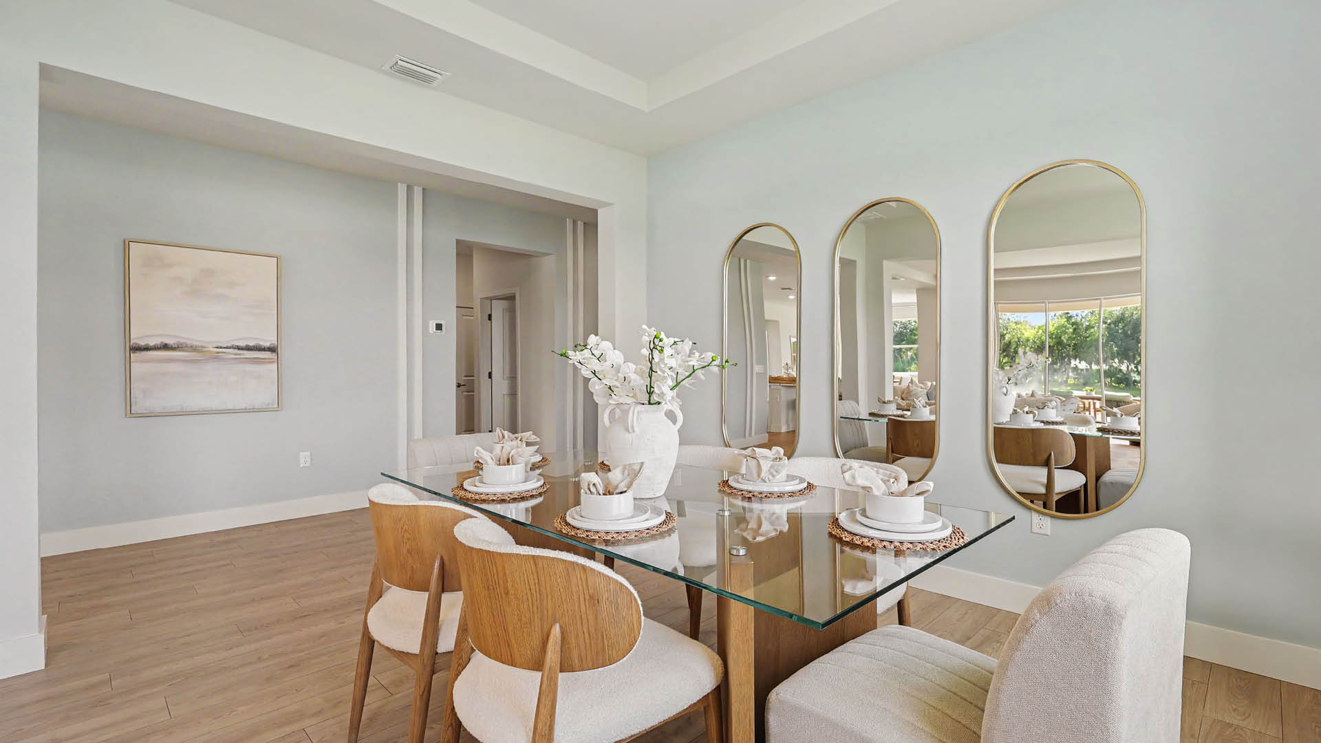 A modern dining room features a glass table set with white dishes and a vase of white flowers, surrounded by wooden chairs with white cushions and a cushioned bench, with three tall oval mirrors on the wall reflecting an adjacent room.