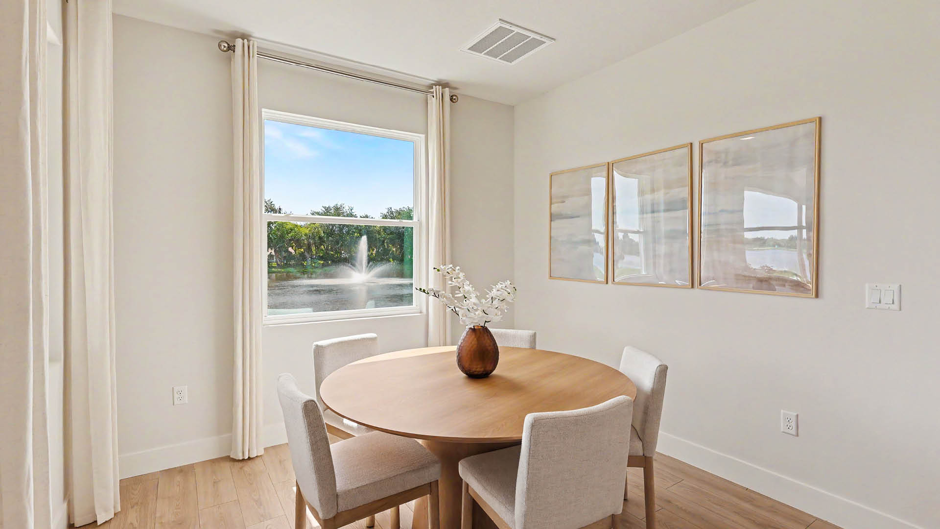 A bright dining area features a round wooden table with a vase of white flowers, surrounded by four upholstered chairs, with a large window showing a fountain outside and three abstract framed artworks on the wall.