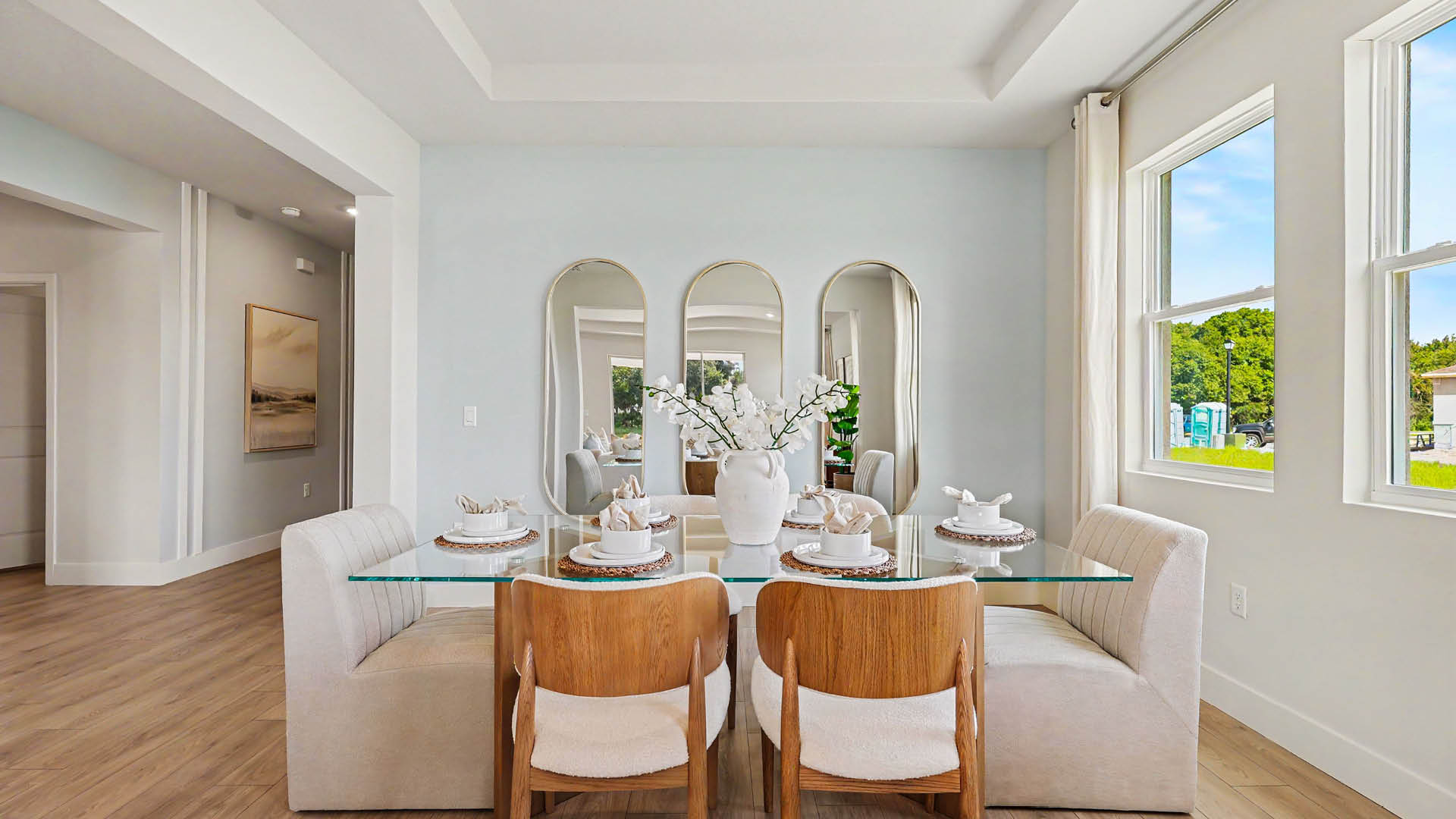 A modern dining area features a glass table set with plates and a white vase with flowers, surrounded by cushioned bench seating and wooden chairs, with three tall mirrors on the wall and large windows letting in natural light.