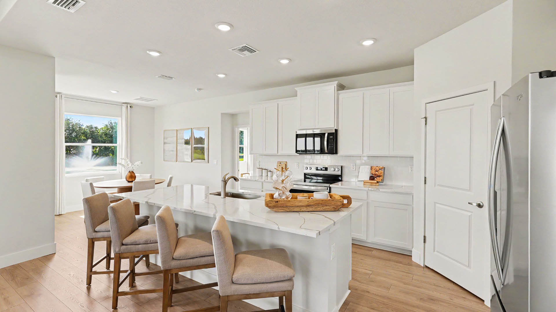 A modern kitchen with white cabinets, an island with four cushioned stools, stainless steel appliances, and a dining table near a window with natural light.