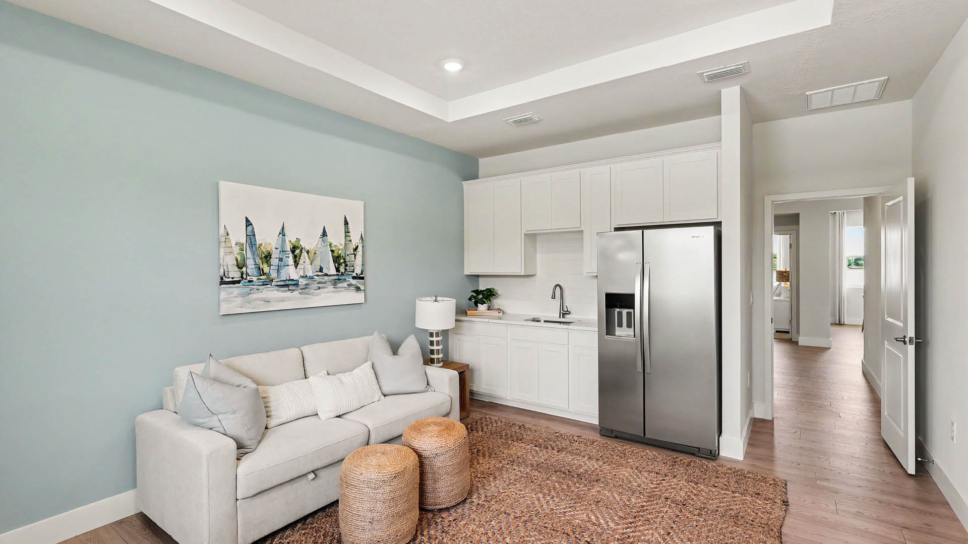 A modern living space features a light gray sofa with pillows, two woven ottomans, a blue accent wall with a sailboat painting, and a compact kitchen area with white cabinets and a stainless-steel refrigerator.