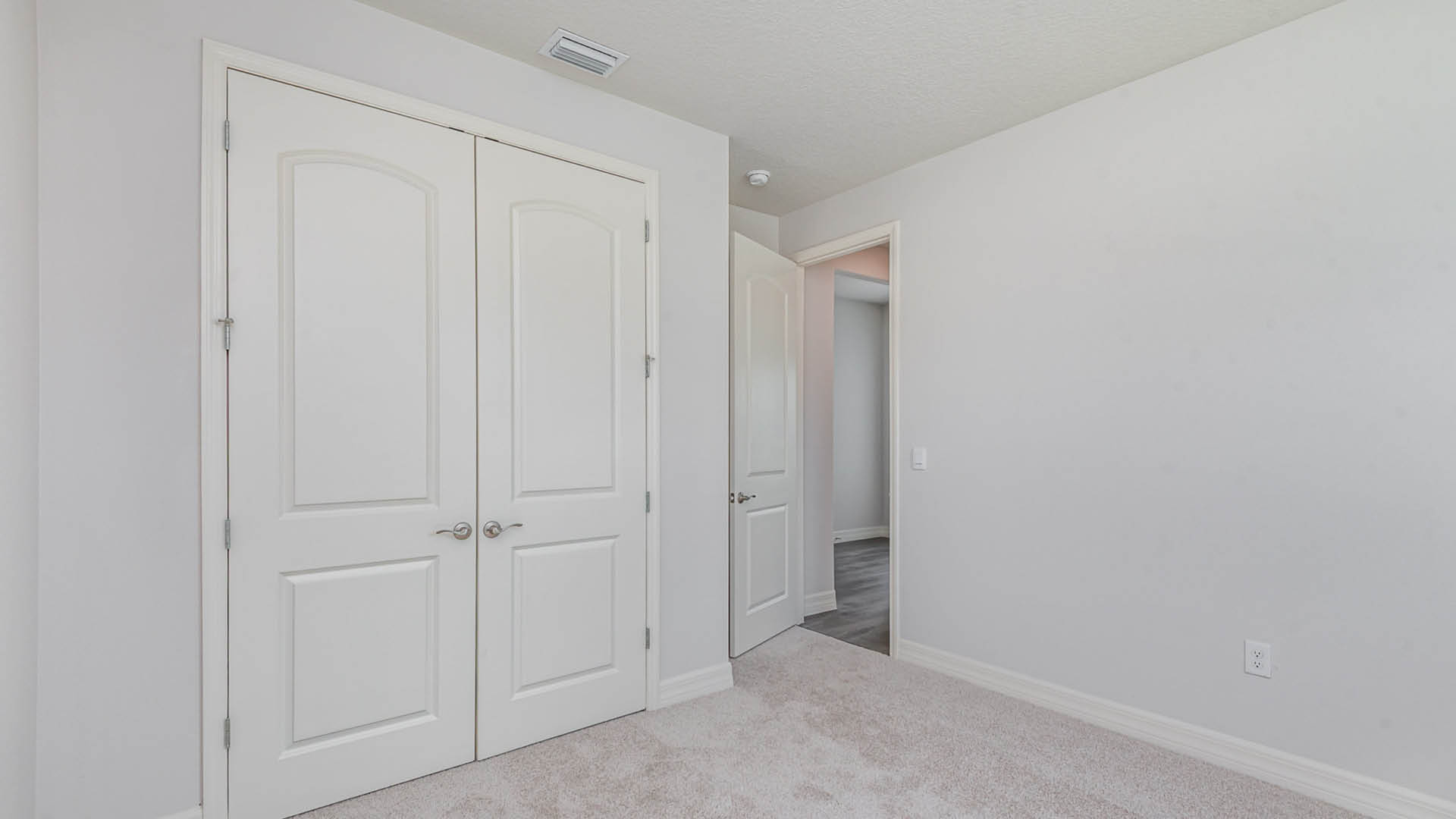 Two white double doors leading to a room, framed by light gray walls and soft carpet, with an open door revealing a hallway beyond.