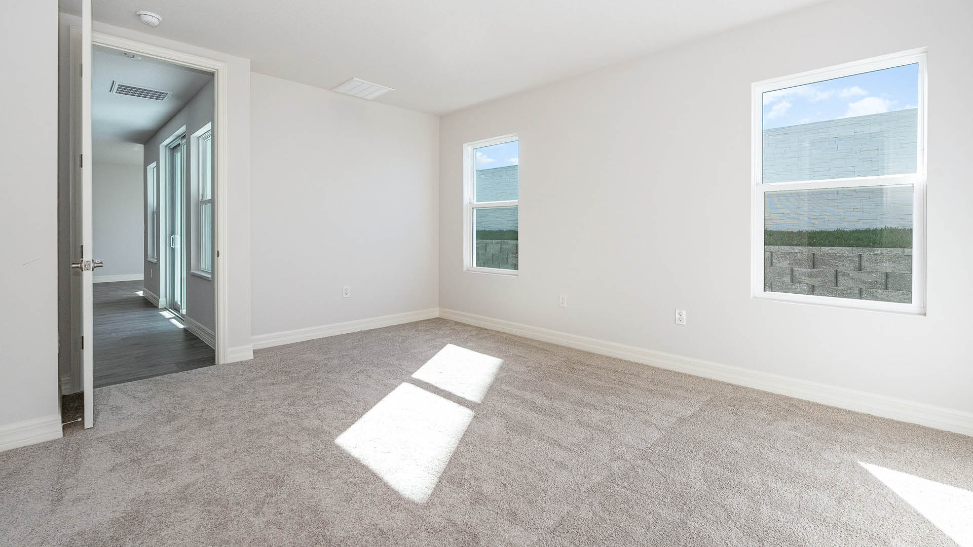 A large bedroom with beige carpet, white walls, and two windows, allowing natural light to illuminate the space. The door to the room is visible, showing the space beyond.