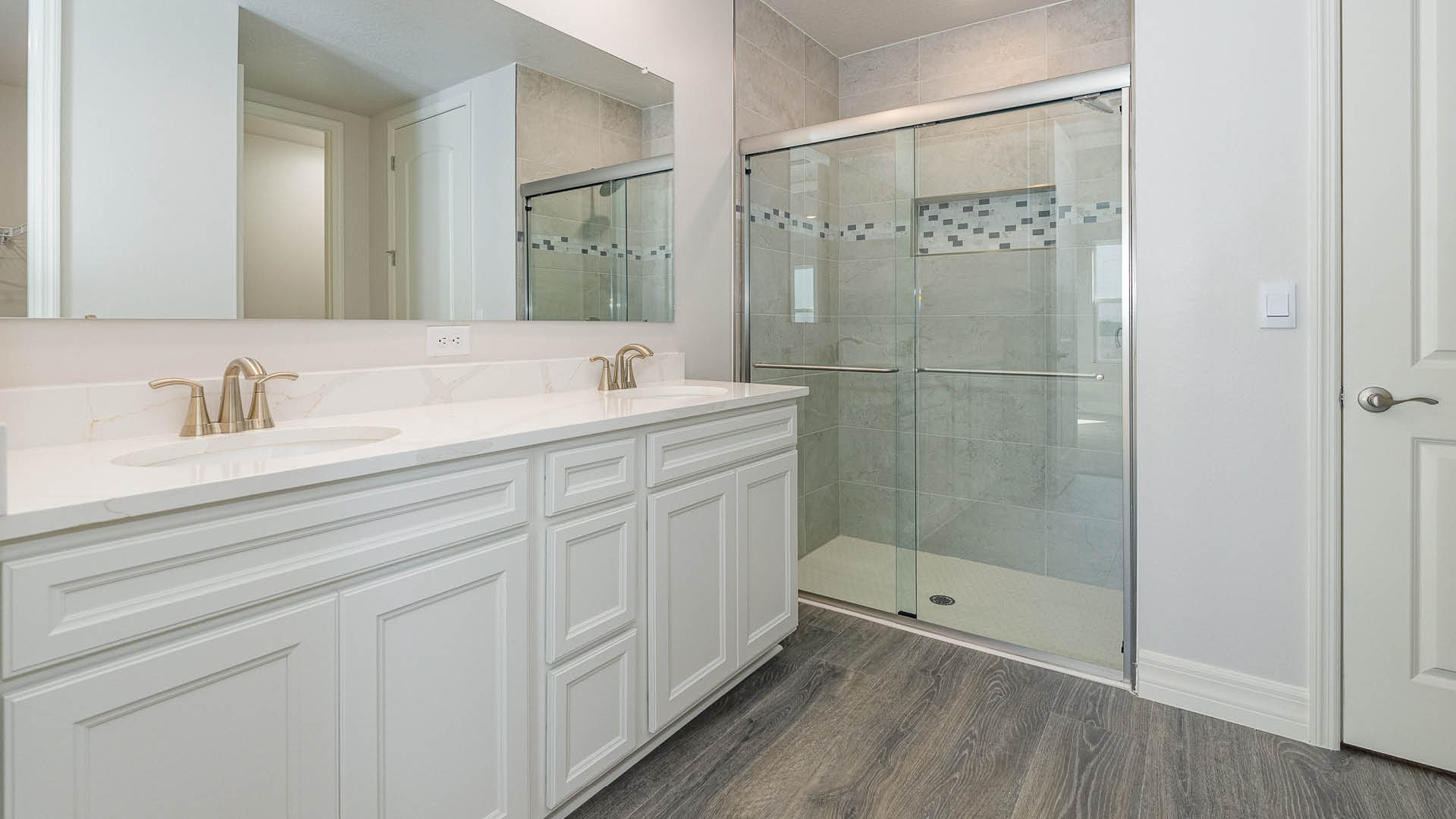 A spacious, ensuite bathroom featuring a double sink vanity with quartz countertops and a walk-in shower. A closed door is visible.