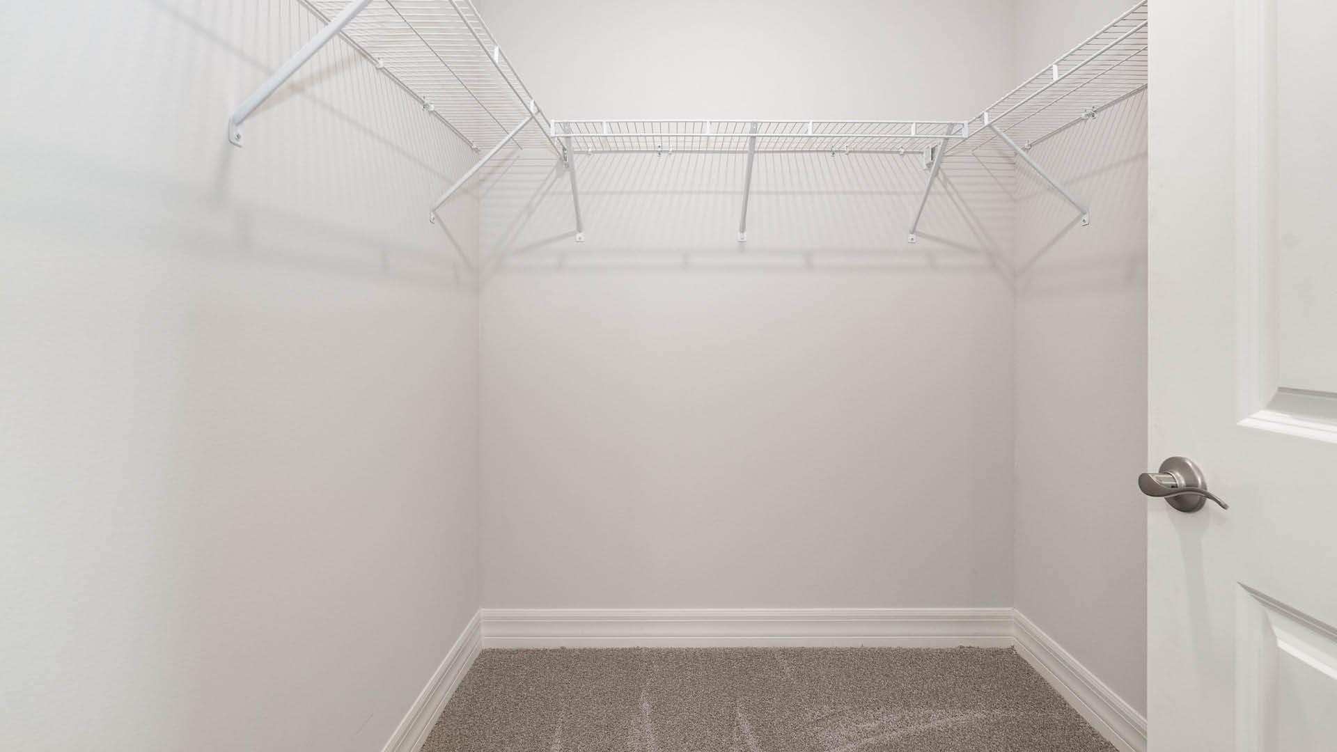 A spacious, walk-in closet with beige carpet, featuring white wire shelves against a light-colored wall.