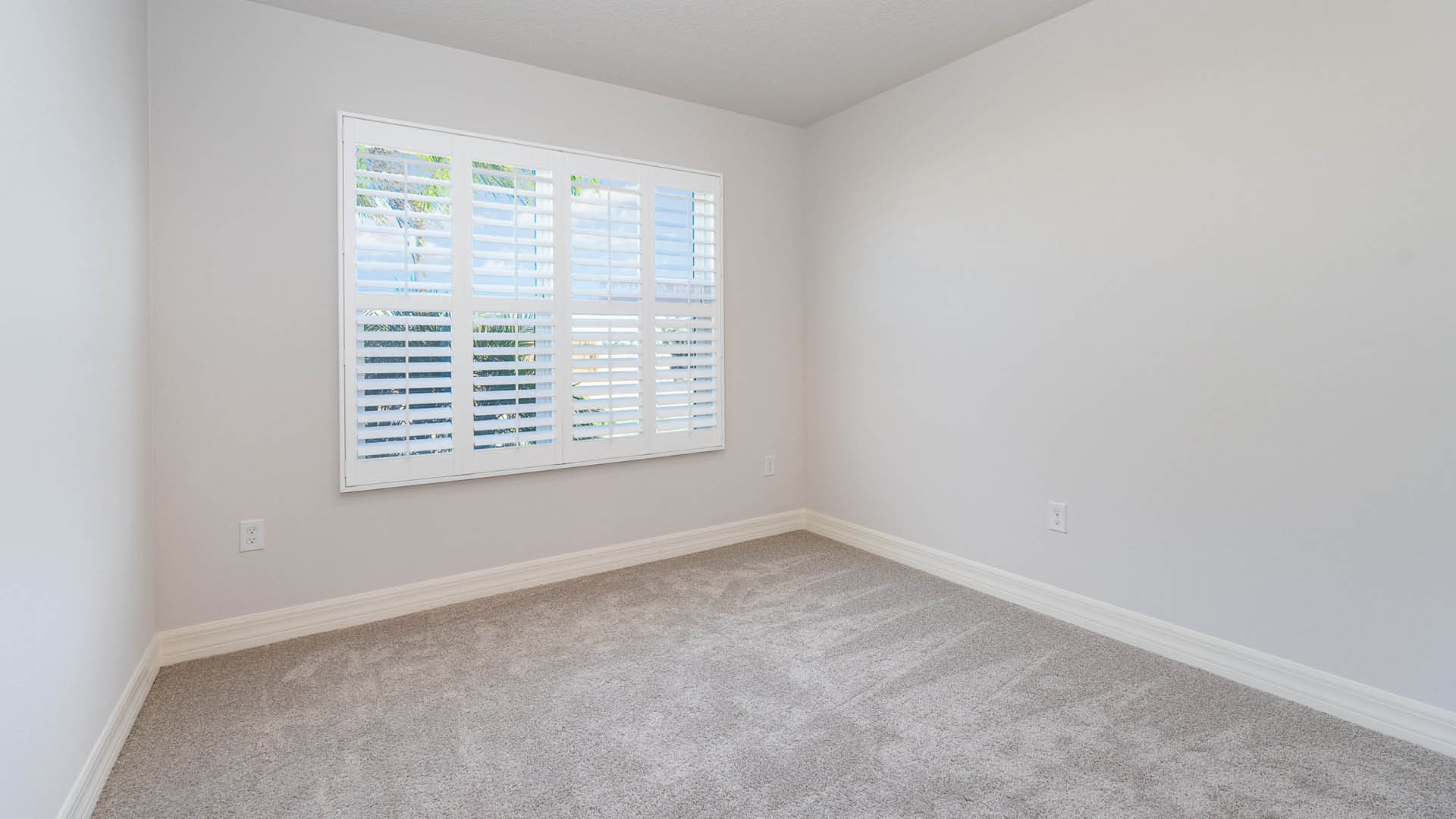 An empty room with beige carpet, white walls, and a window with blinds, allowing natural light to illuminate the space.