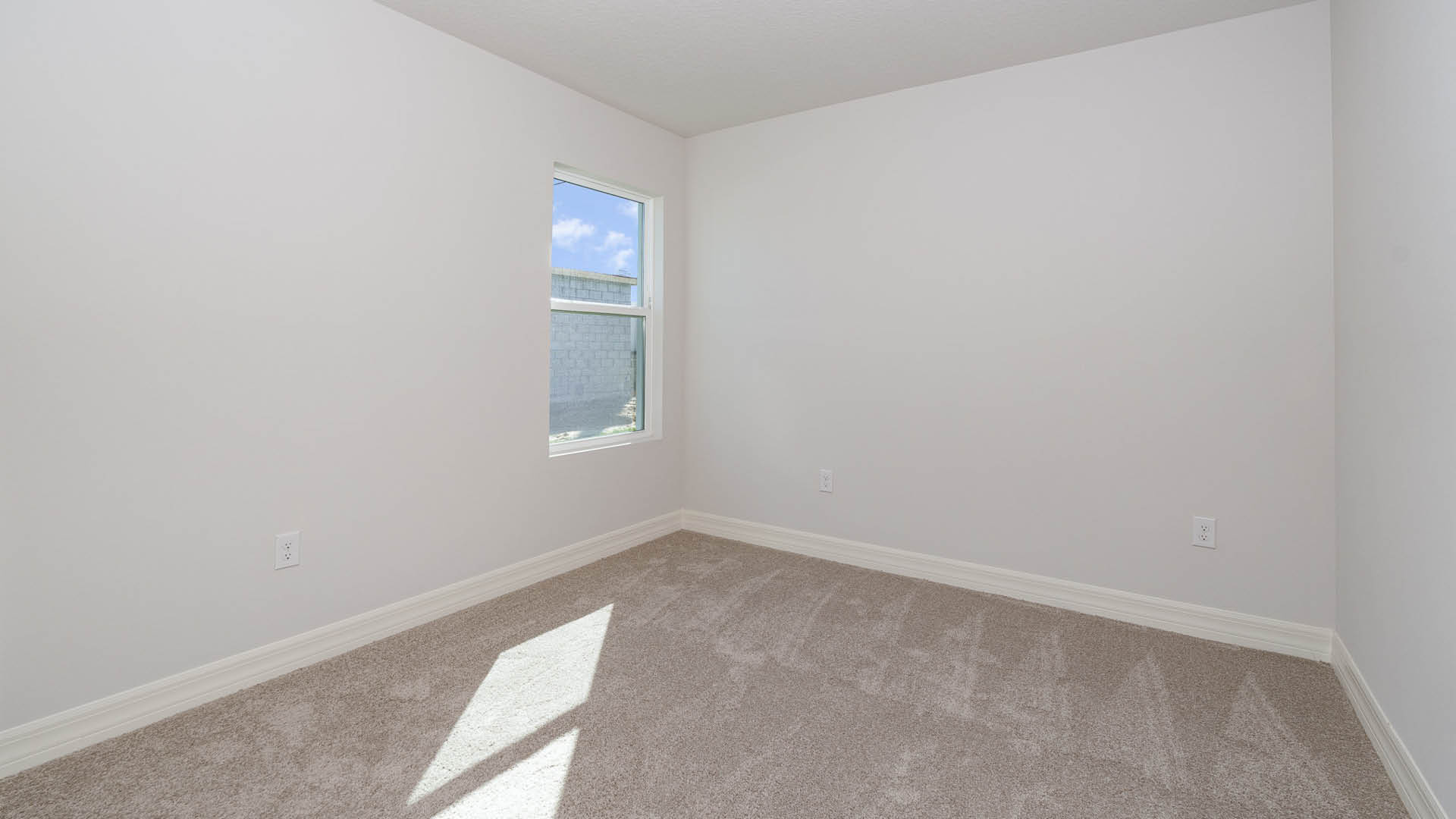 An empty room with beige carpet, white walls, and a window, allowing natural light to illuminate the space.
