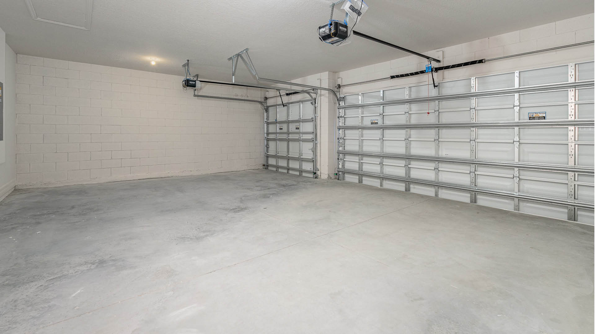 A spacious, 3-car garage with concrete flooring, two garage doors, and a white cinder block wall. A ceiling access panel is visible.