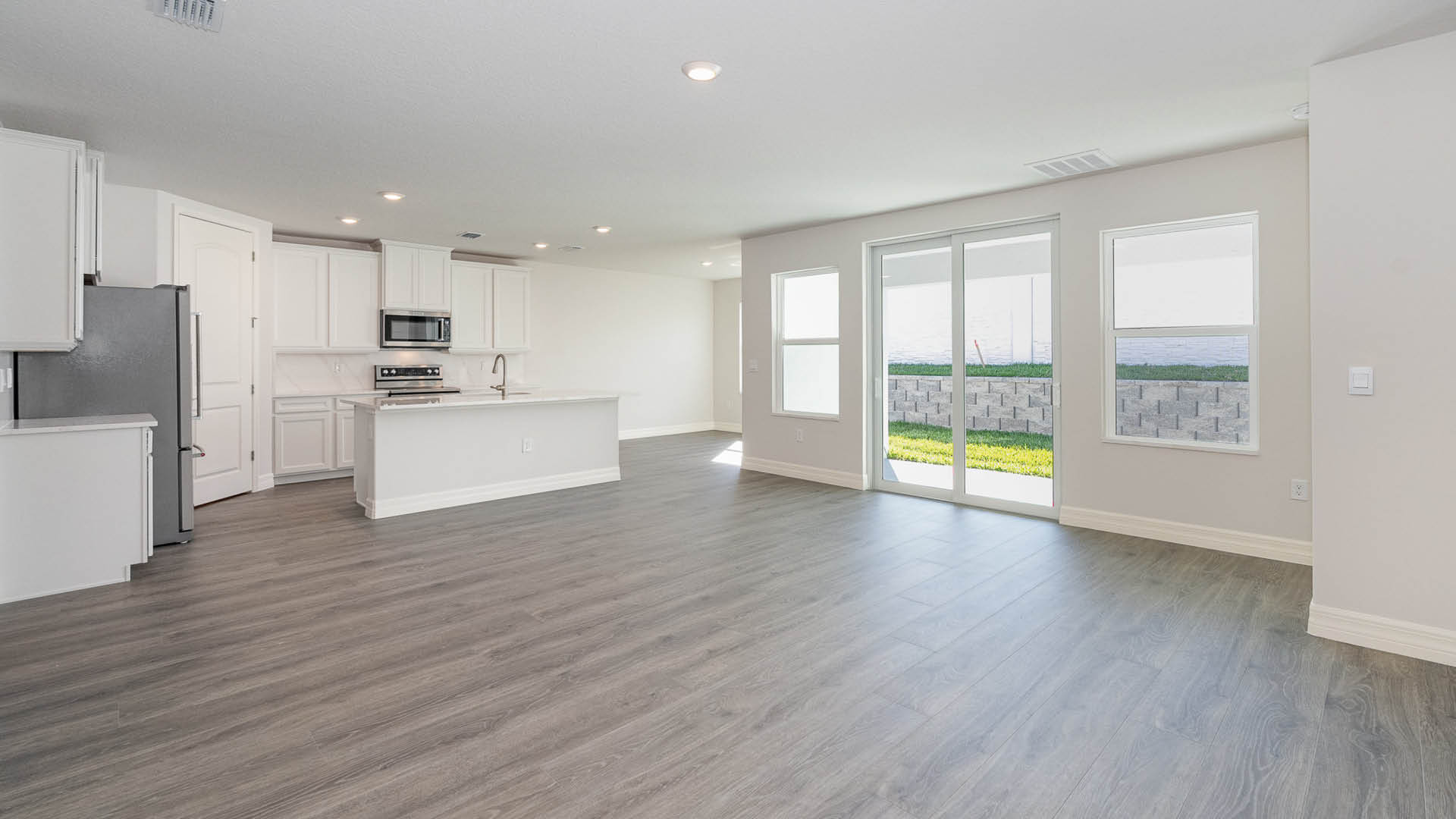 A spacious room, with a view of a kitchen and dining area and Mohawk® Revwood Select Plank Flooring throughout. There are large windows and sliding glass doors leading to a covered patio.