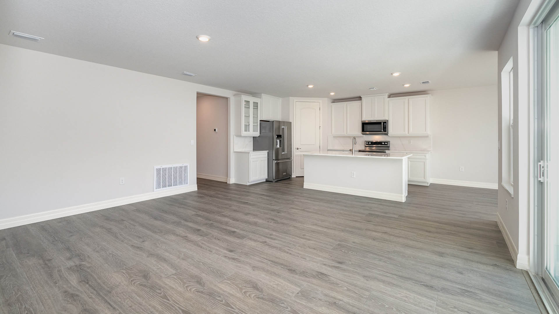 A spacious room, with a view of a kitchen area and Mohawk® Revwood Select Plank Flooring throughout. A hallway leading elsewhere is visible.