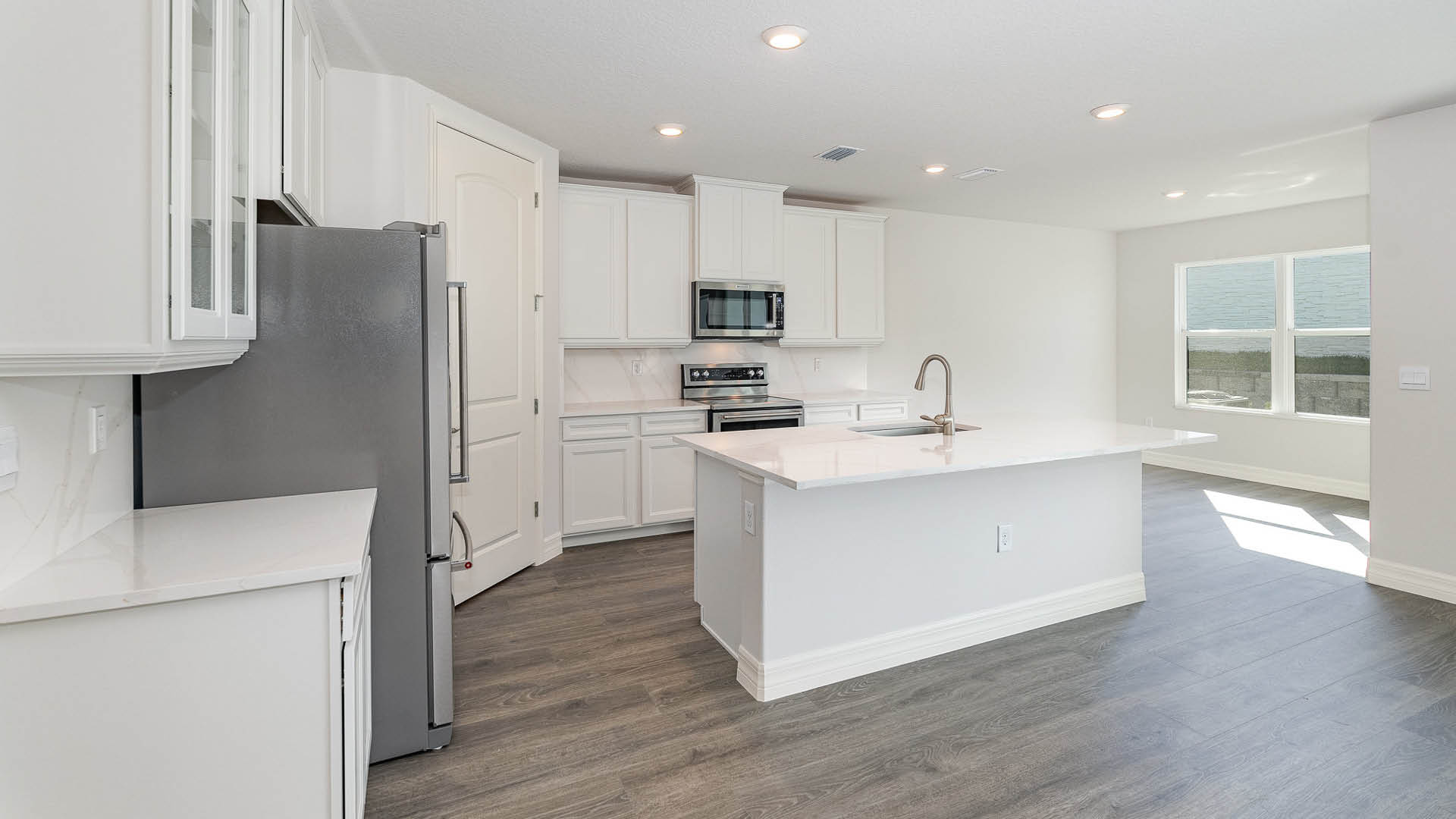 A large kitchen with quartz countertops, stainless steel appliances, and an island with room for seating. A dining area featuring a large window is visible.
