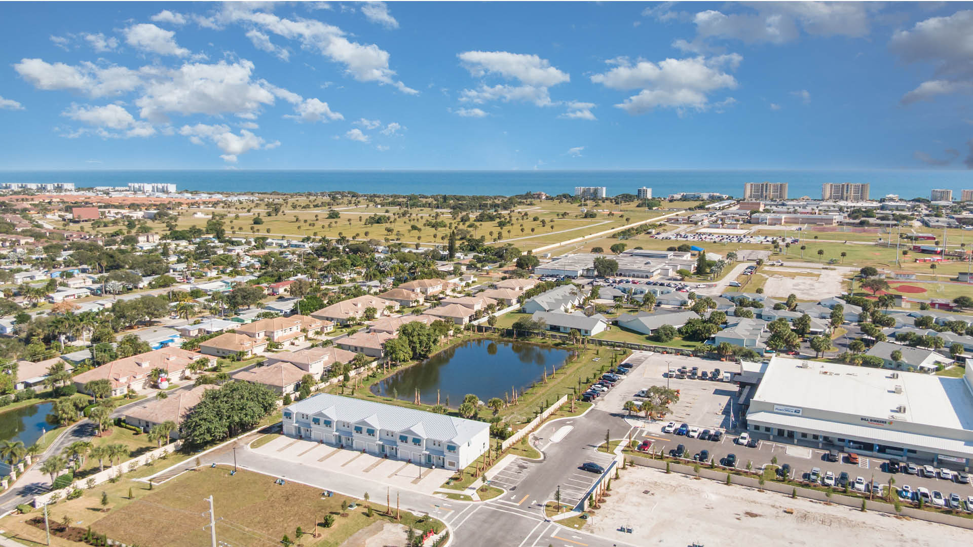 Aerial view of a Tortuga Cay featuring townhomes, a pond, commercial areas, and the ocean in the distance under a blue sky.