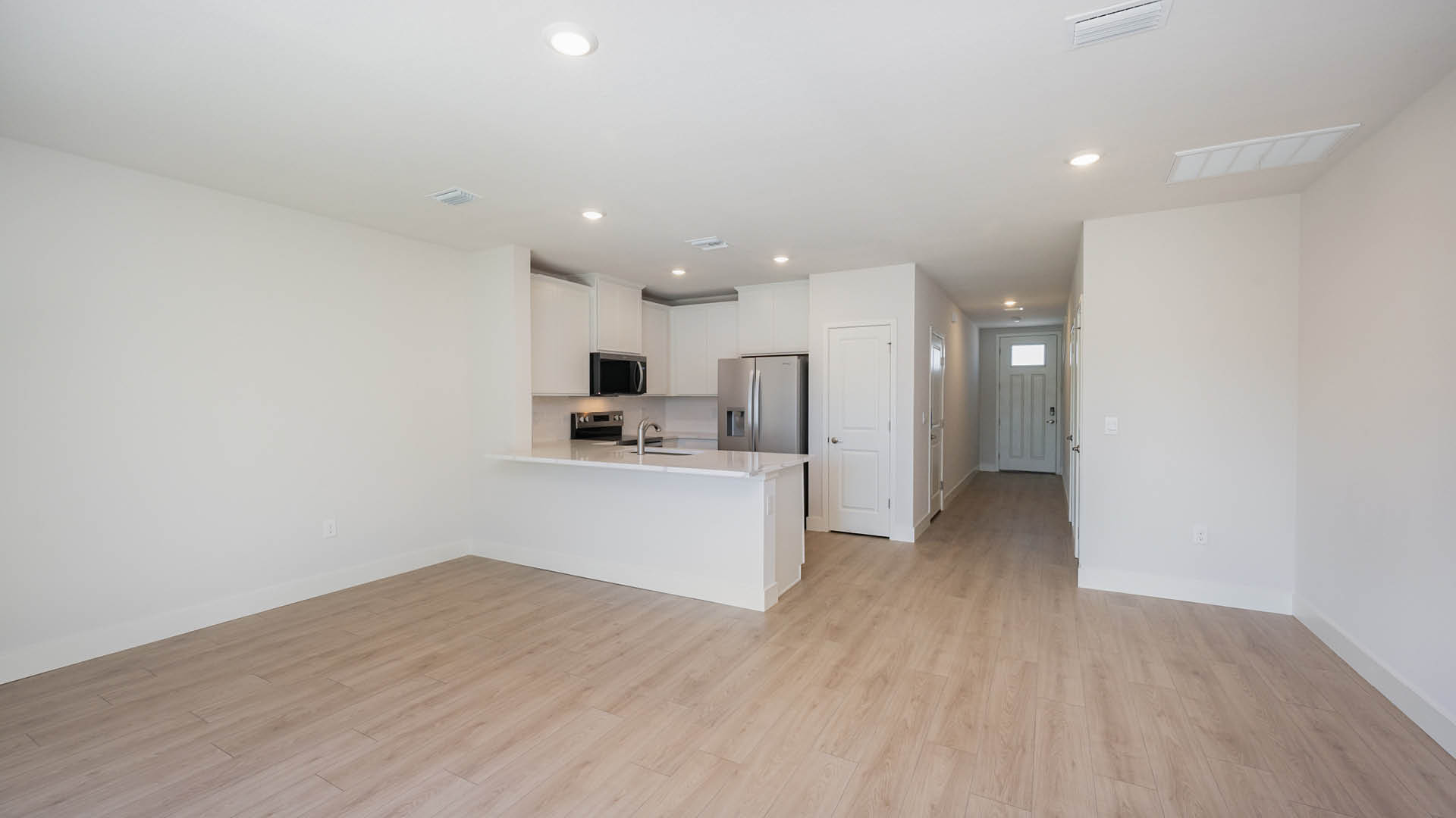 Spacious, light-filled living area with light-colored walls and modern wood-style flooring leading to a contemporary kitchen.