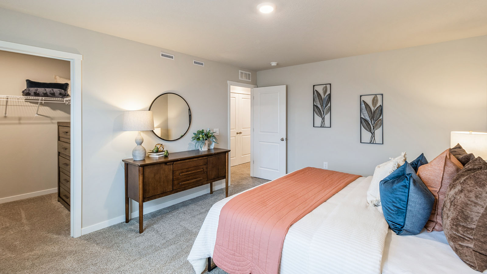 A modern bedroom with a neatly made bed, warm-toned bedding, a wooden dresser with a round mirror, and an open walk-in closet with built-in shelving.