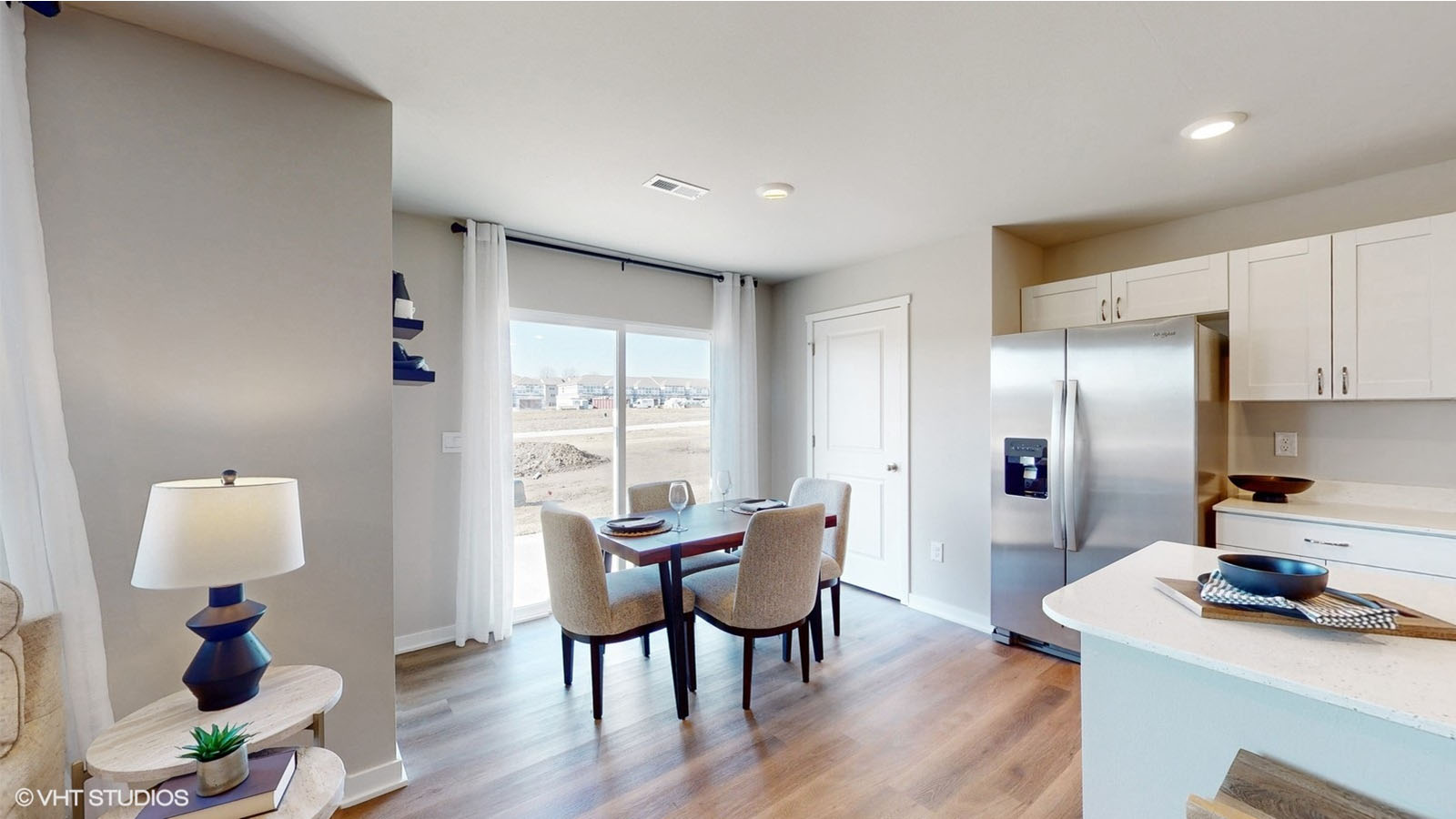 An open-concept kitchen and dining area featuring white cabinetry, stainless steel appliances, and a cozy dining nook with a wood table and four upholstered chairs, all illuminated by natural light from sliding glass doors with a view of the backyard.