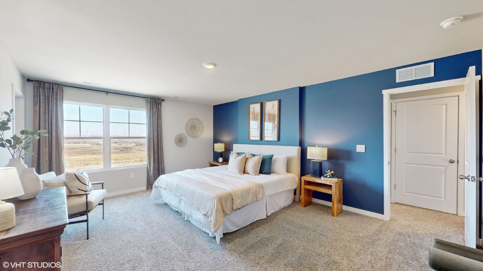 A bright and airy primary bedroom with a striking navy accent wall behind a king-sized bed, large windows that flood the space with natural light, plush carpet flooring, and warm wood nightstands.