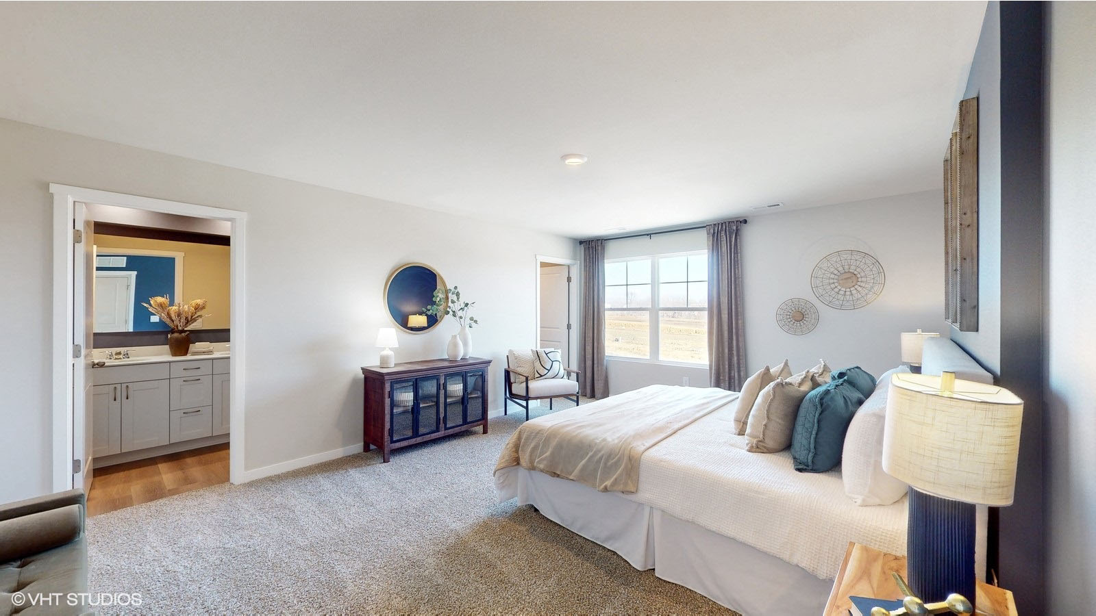 A spacious and sunlit primary bedroom featuring soft neutral walls, a large window with floor-length curtains, and cozy carpeted floors; the room connects to an en suite bathroom and includes stylish decor like a round navy mirror, wood dresser, and coordinating blue and beige accents.