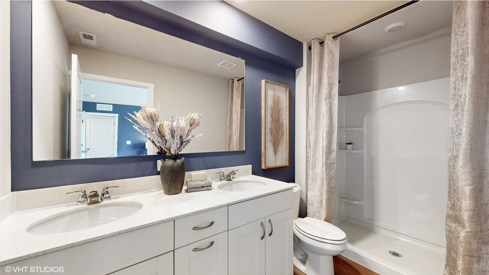 A modern bathroom with a navy accent wall above a wide mirror, double-sink white vanity, and a glass vase filled with dried botanicals, featuring a walk-in shower with built-in shelving and soft neutral-toned curtains.
