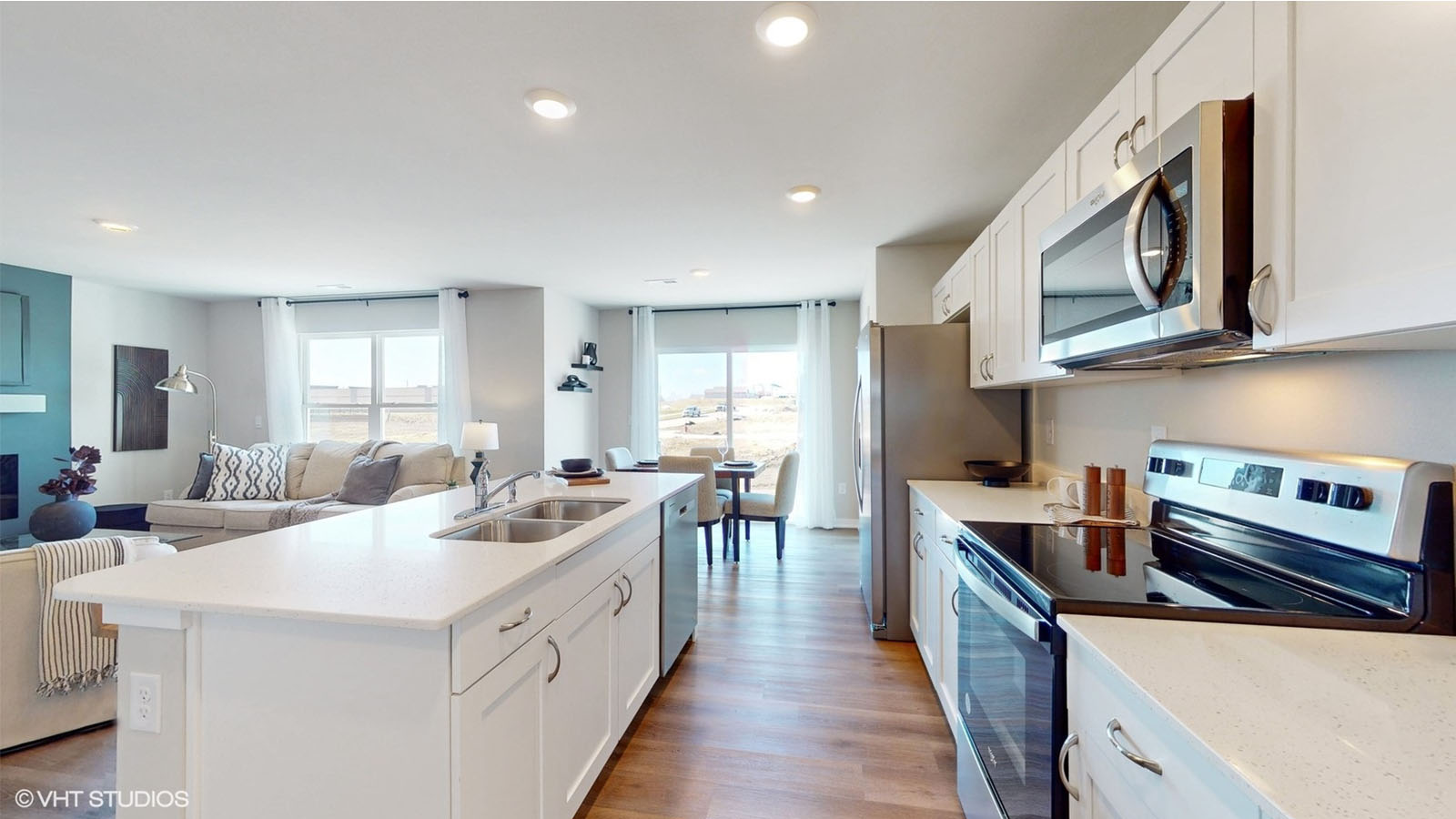 A bright, open-concept kitchen and living area featuring sleek white cabinetry, stainless steel appliances, and a spacious island with an integrated sink and dishwasher, seamlessly flowing into a sunlit dining space and cozy living room with modern furnishings and floor-to-ceiling windows.