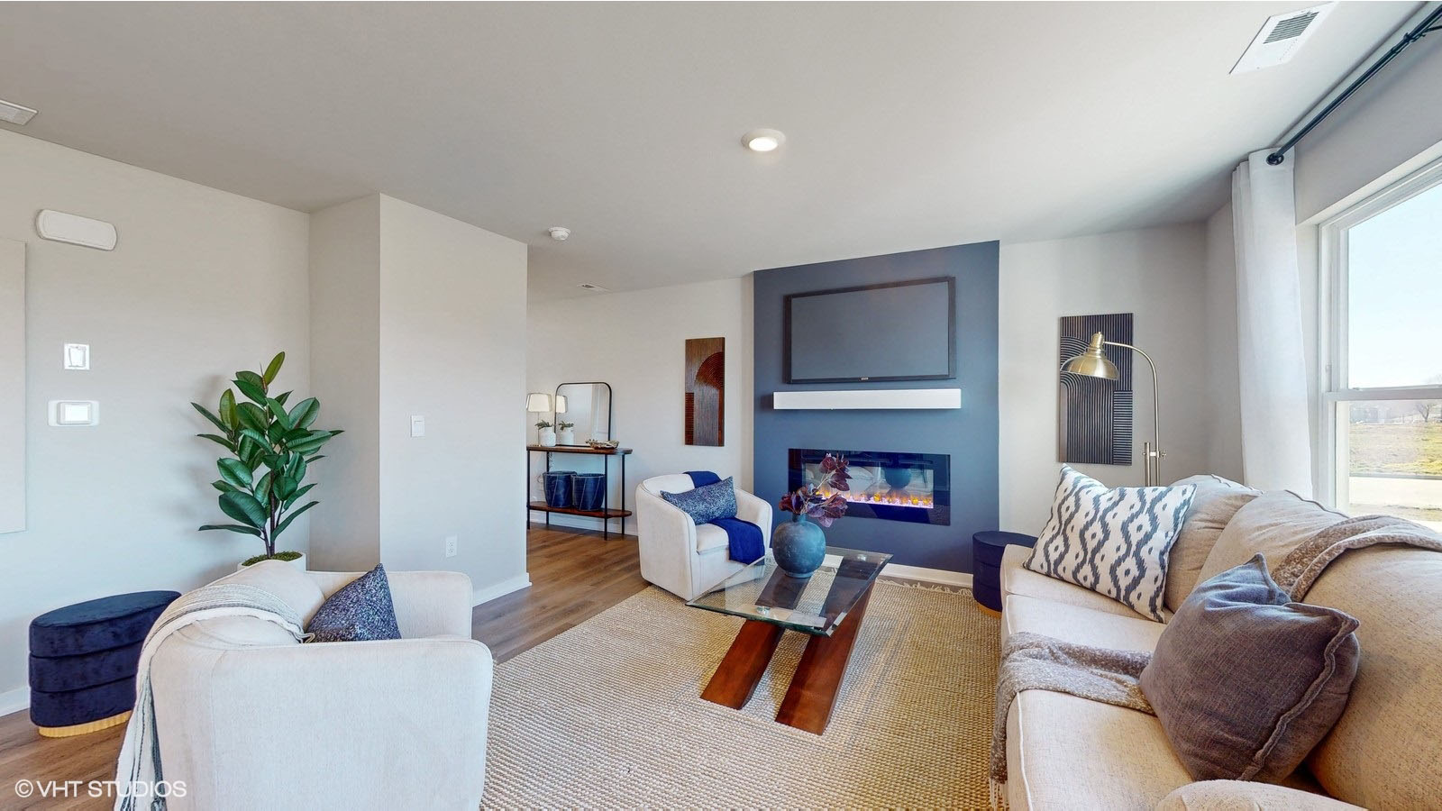 A contemporary living room showcasing a neutral-toned sectional sofa and armchairs, centered around a dark accent wall with a mounted TV and modern electric fireplace, with wood-look flooring, a glass coffee table, and a large window filling the space with natural light.